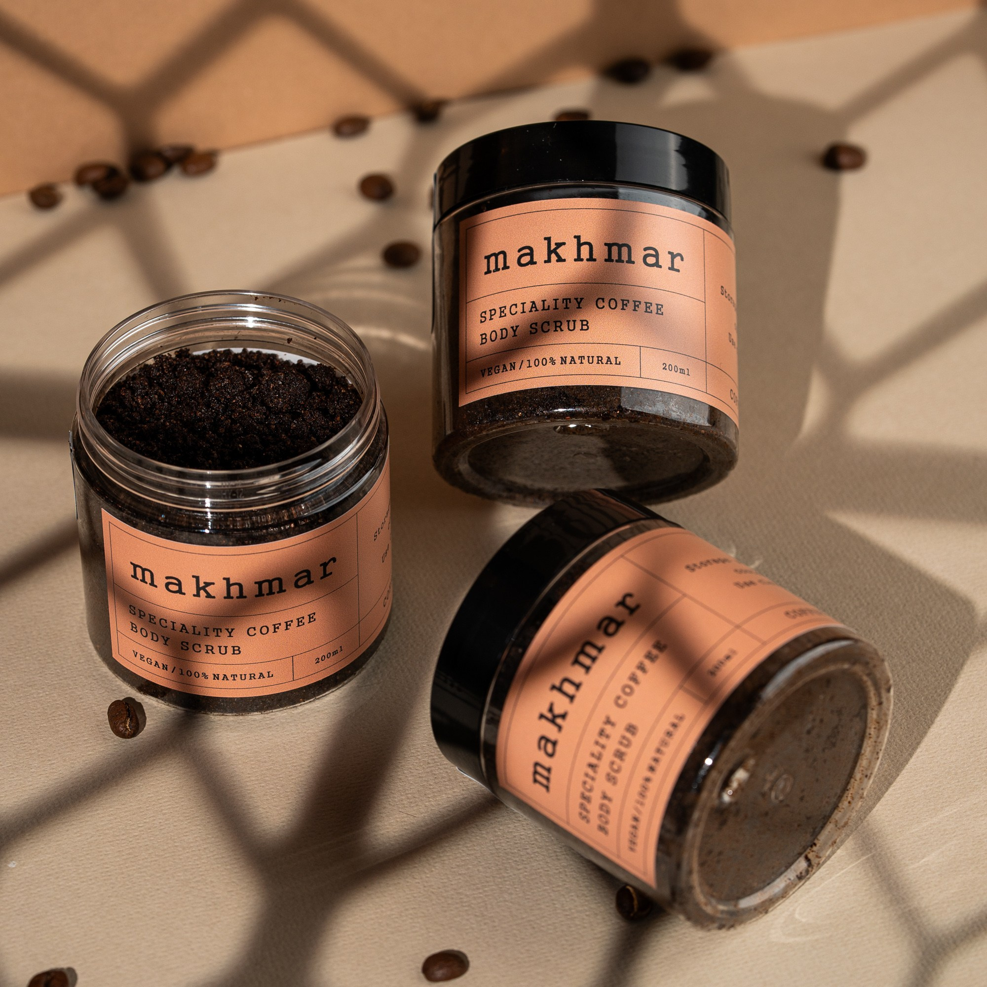 Makhmar Speciality Coffee Body Scrub üç qabda, qəhvə dənələri və kölgə effektli minimalistik məhsul fotoşəkili.