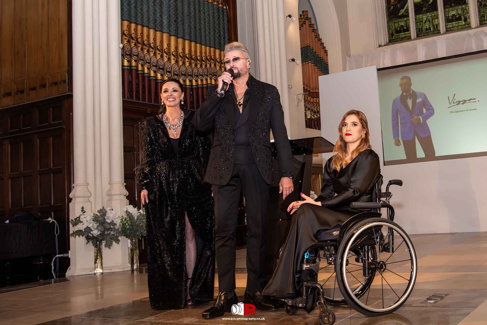 Presenter with a microphone standing alongside a seated female model in a wheelchair and a glamorous co-host in a sparkling black gown.