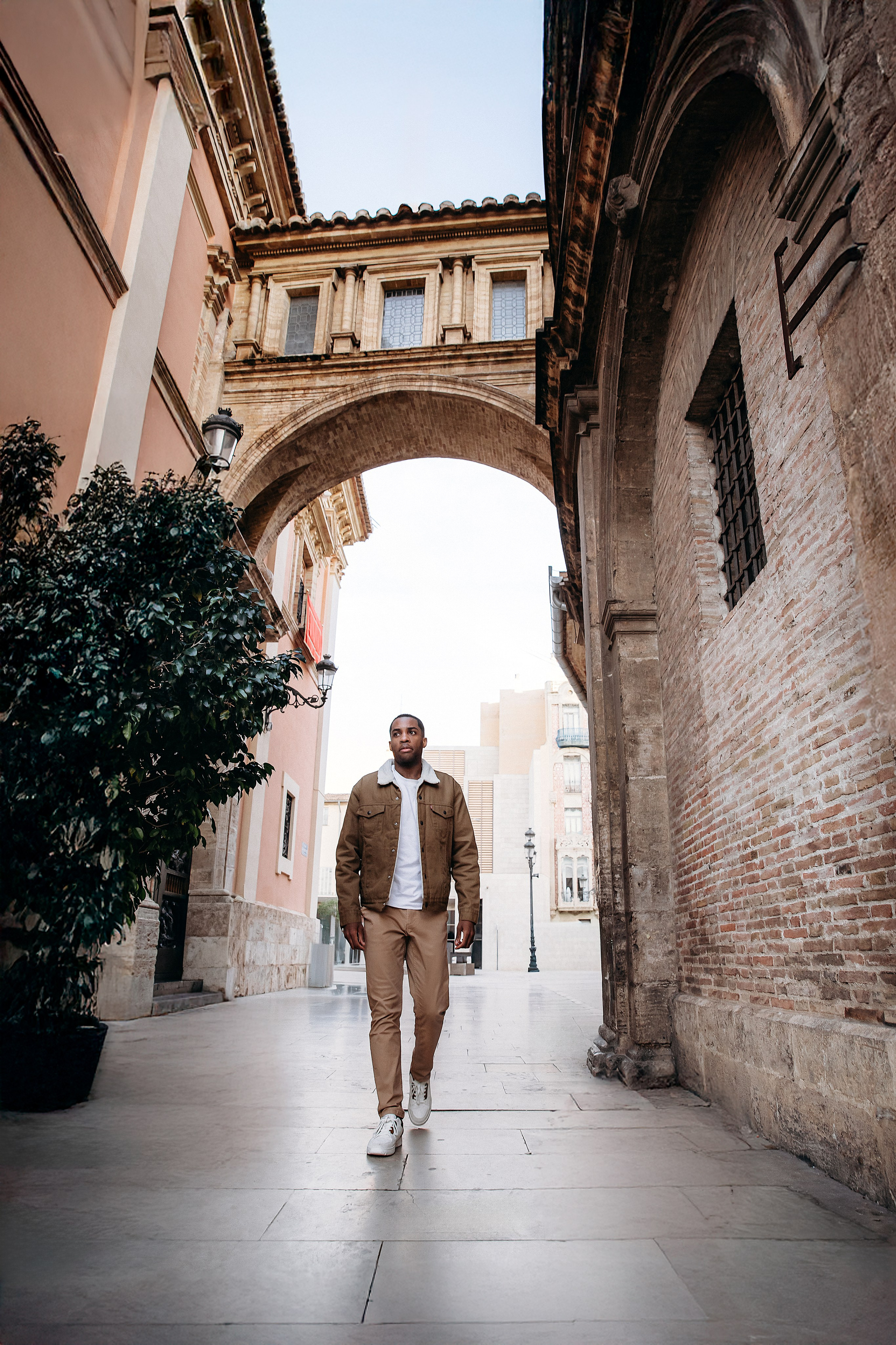 Мужская портретная фотосессия в Валенсии, Испания, с уверенным мужчиной, проходящим через историческую каменную арку в старом городе, в повседневной куртке и светлом образе, снятым при естественном дневном свете с элегантной средиземноморской архитектурой и городской атмосферой — идеально для мужской портретной фотографии, lifestyle-фотосессий для мужчин, портретов для персонального бренда, городских fashion-сессий и профессиональных мужских фотосессий в Валенсии и по всей Испании.
