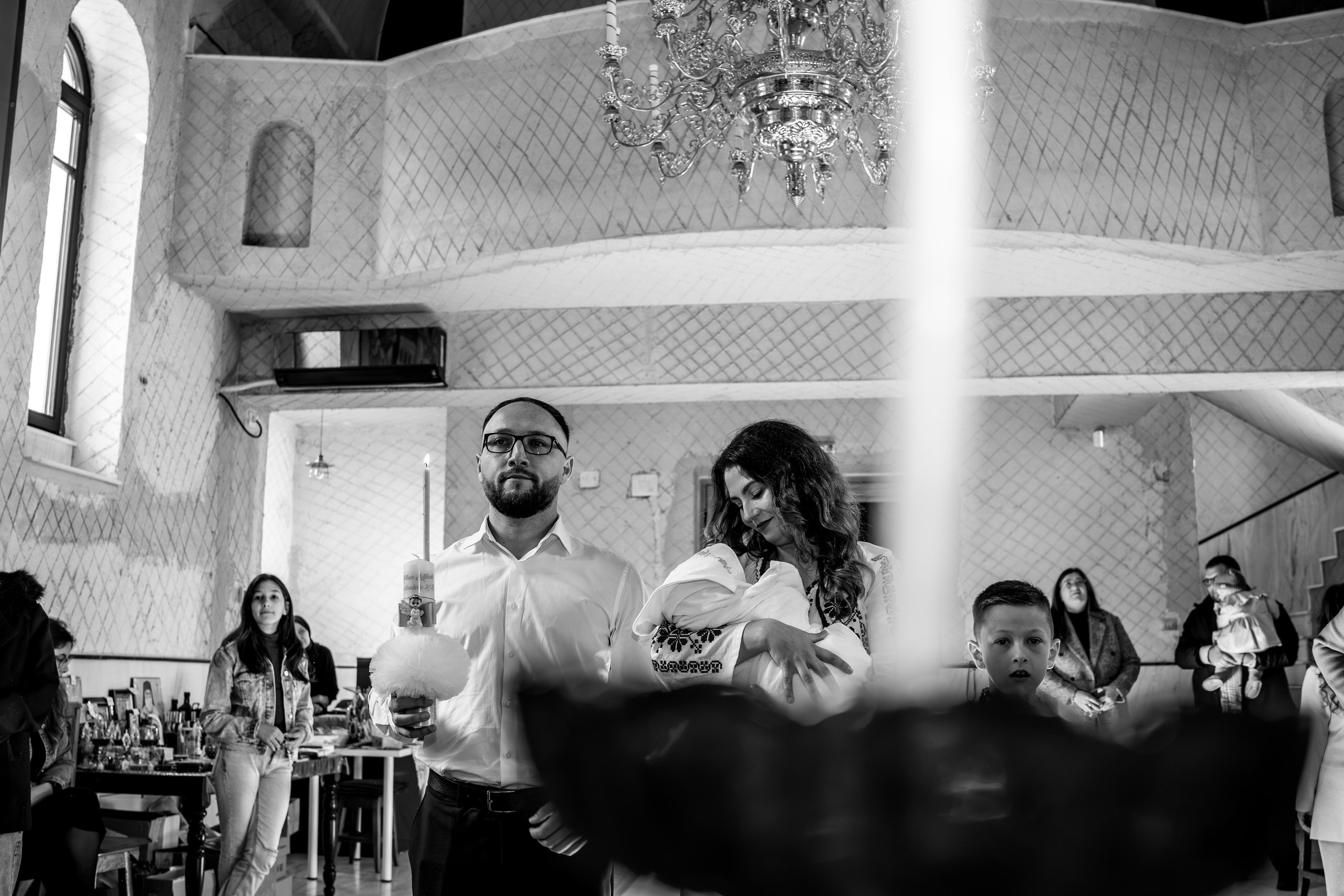 Fotografie alb-negru panoramică după botezul lui Arthur Mihail. Nașul ține în mână lumânarea de botez aprinsă, iar nașa, ținând pruncul, îl privește cu o căldură și o grijă profunde. Un cadru intim și puternic, plin de simbolism și promisiune.