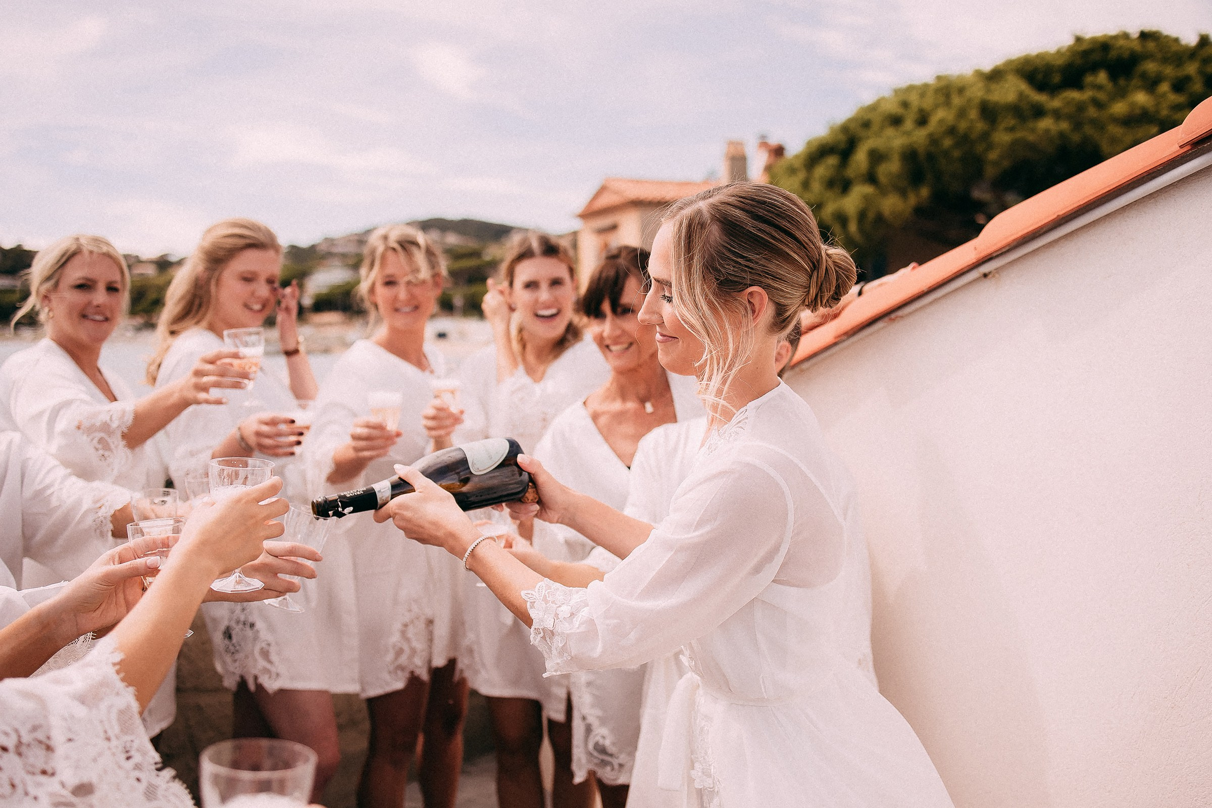 A candid shot of bridesmaids cheering as the bride pours champagne on a sunny terrace overlooking the tranquil landscape of Provence.