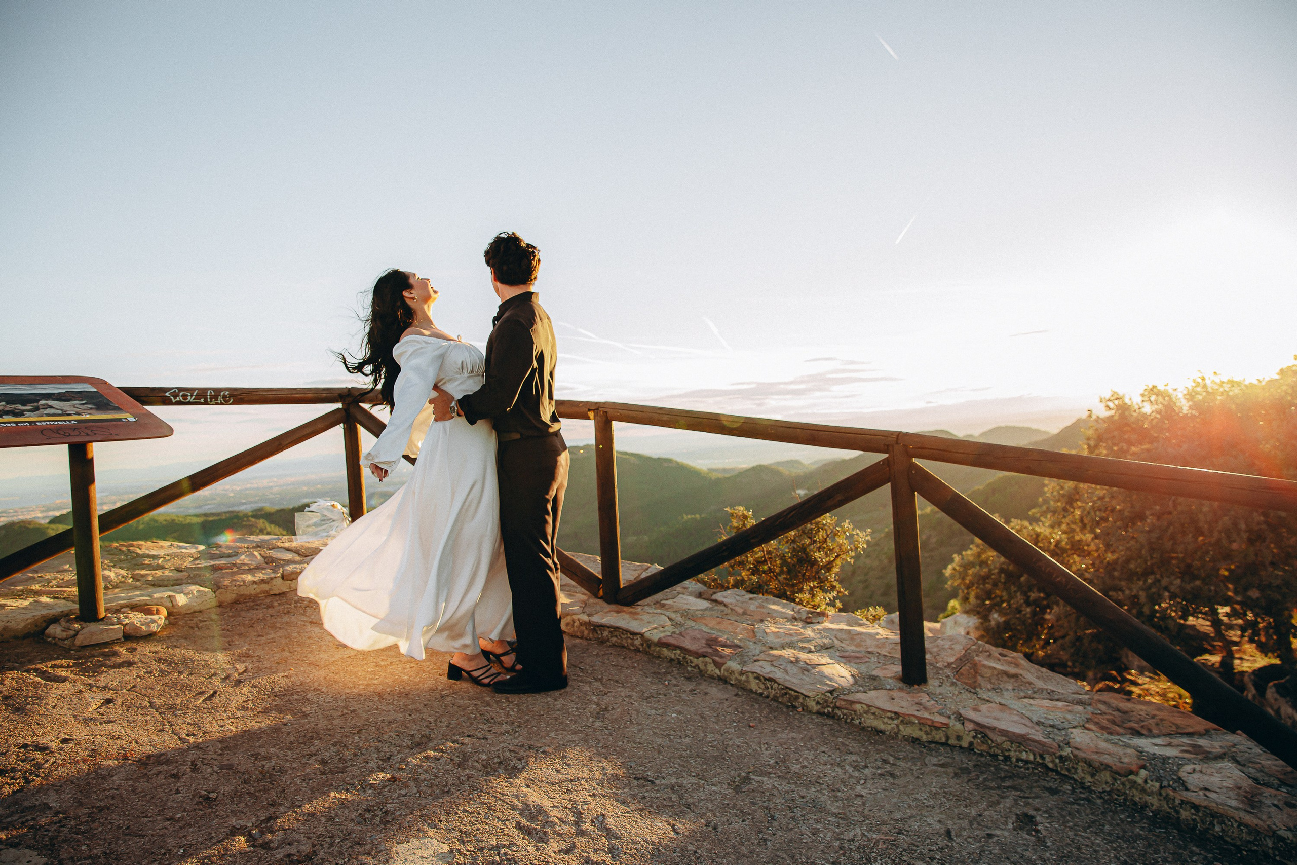 Engagement photoshoot in València, Spain, featuring a couple embracing on a scenic mountain viewpoint with wooden railings at sunset, the bride in a flowing white dress and the groom in dark attire, capturing a romantic and cinematic wedding love story — ideal for engagement photography, pre-wedding sessions, elopements, and professional engagement and wedding photoshoots in València and across Spain.