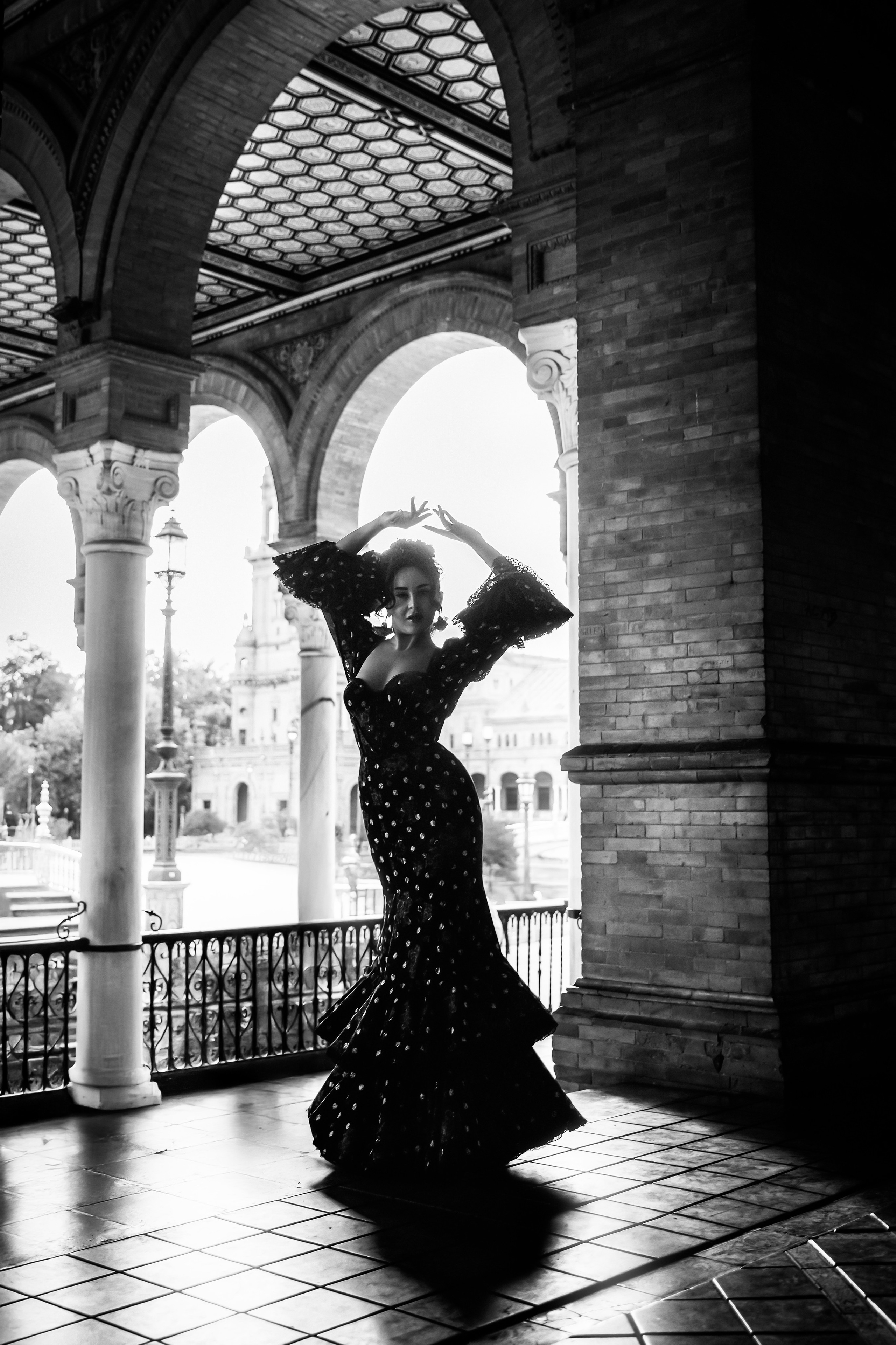 Драматическая фотосессия в стиле фламенко в Севилье, Испания — женщина в платье в горошек исполняет традиционный танцевальный жест под арками площади Испании. Съёмка демонстрирует архитектурную красоту и культурную элегантность Испании, идеально для художественной портретной фотографии в Севилье.