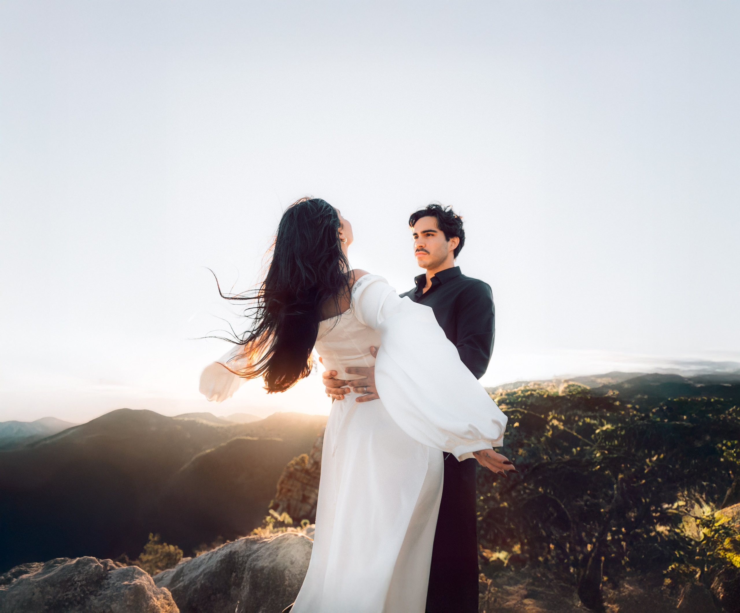 Engagement photoshoot in València, Spain, featuring a romantic couple posing on scenic mountain cliffs at golden hour, with the groom holding the bride in a flowing white dress against a vast Mediterranean landscape — an emotional engagement and wedding love story image ideal for engagement photography, pre-wedding sessions, elopements, and professional wedding photoshoots in València and across Spain.