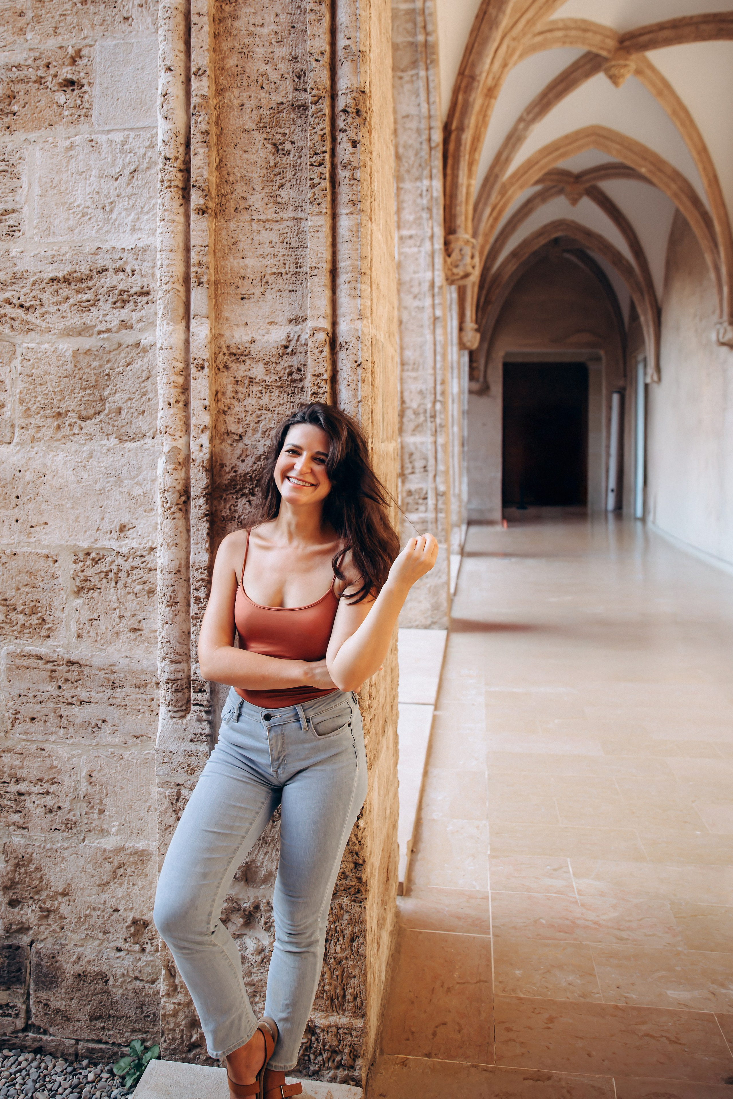 Женская фотосессия в Валенсии, Испания, с улыбающейся женщиной, прислонённой к историческим каменным аркам с готической архитектурой, мягким естественным светом и расслабленной уверенной позой — идеально для женской портретной фотографии, лайфстайл-съёмок, портретов при естественном свете и профессиональных женских фотосессий в Валенсии и по всей Испании.