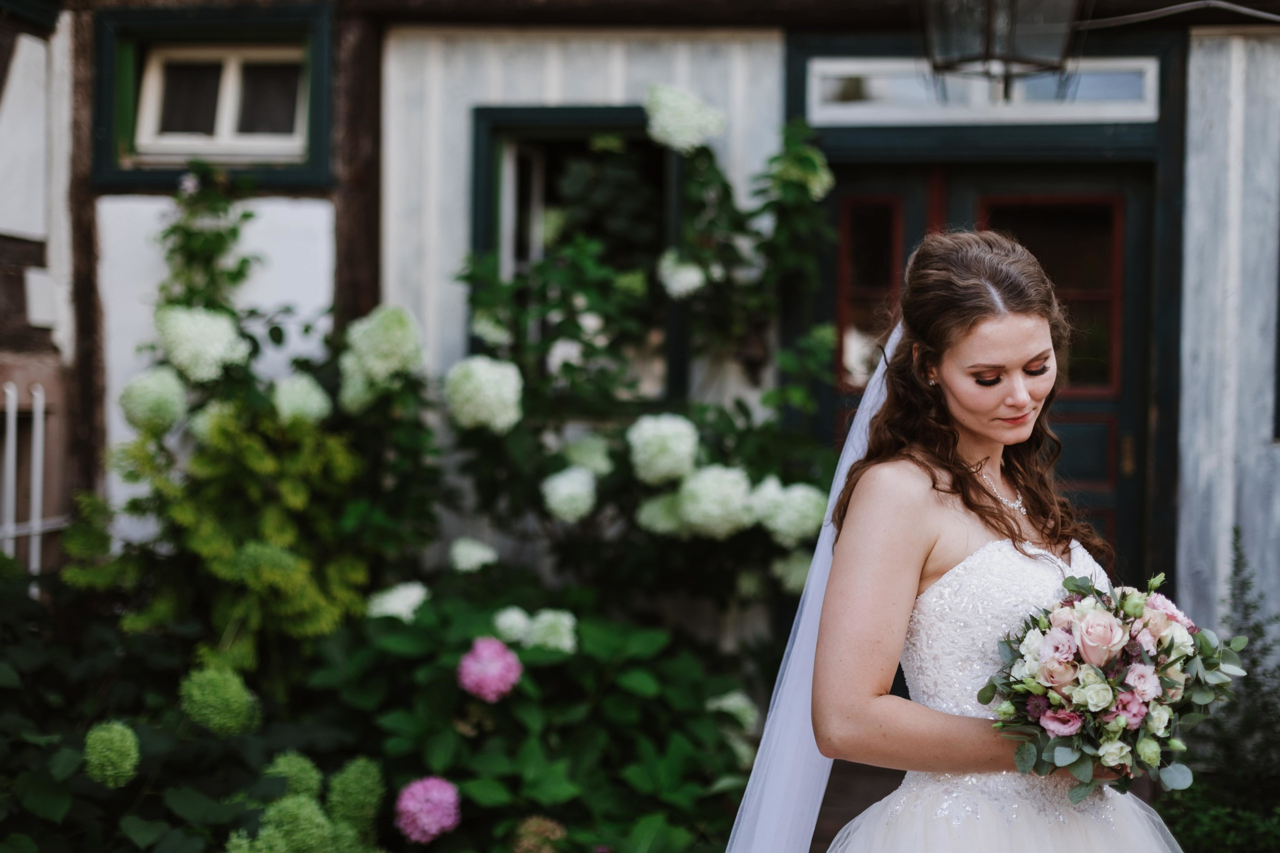 Die Braut betrachtet ihren Brautstrauß und steht vor einem großen Hortensienstrauch.