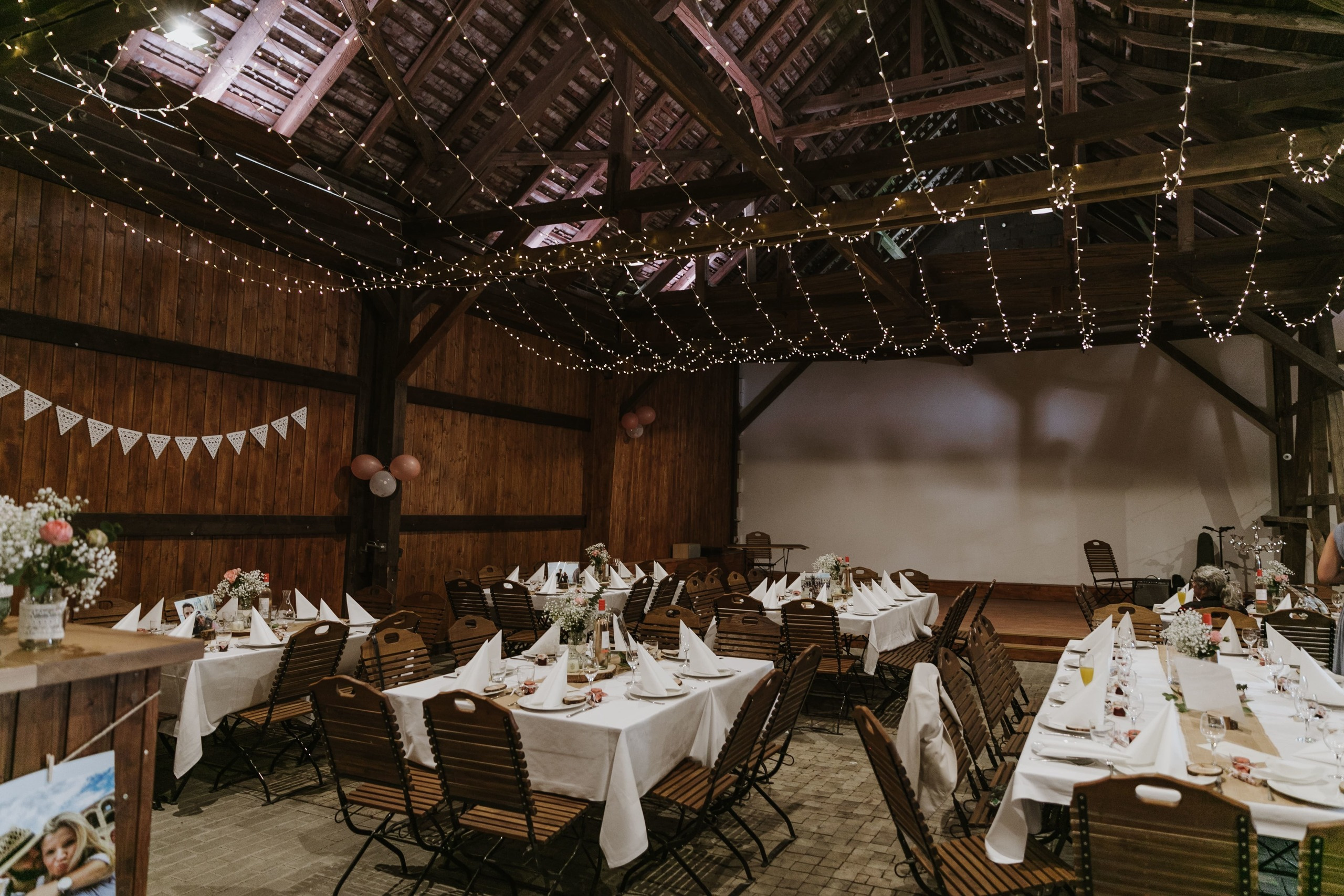 Rustikal dekorierte Hochzeitsscheune mit Lichtergirlanden und Holzmöbeln.