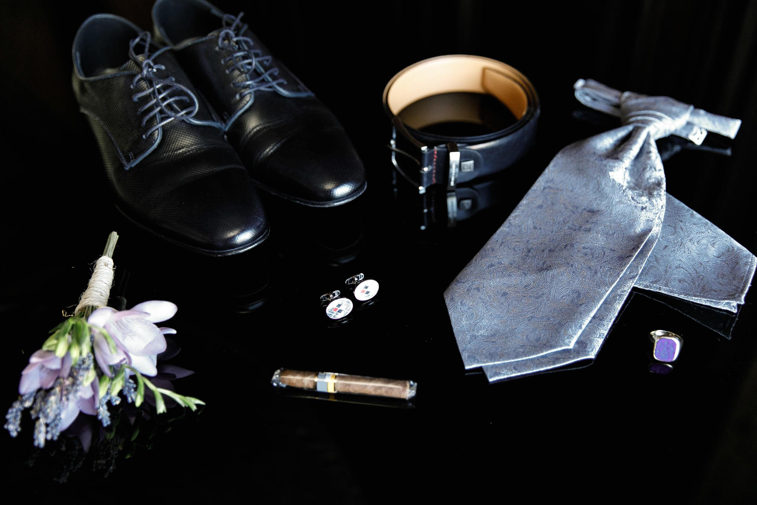 Die Details des Hochzeitsanzugs des Bräutigams: Schuhe, Krawatte, Ansteckblume, Zigarre, Manschettenknöpfe und Ring.