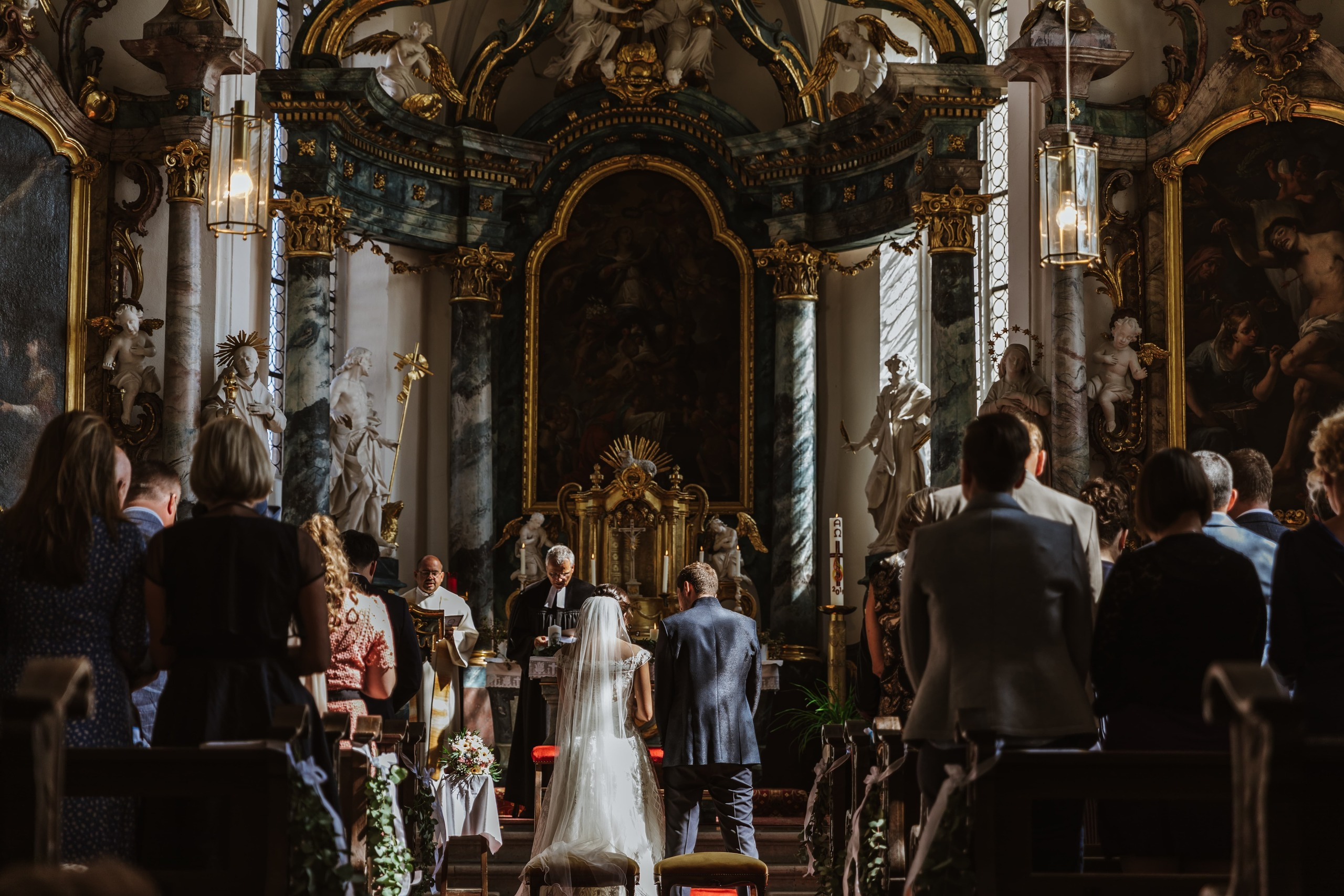 Der Bräutigam, die Braut und die Hochzeitsgäste stehen zum Gebet in einer beeindruckenden barocken Kirche.