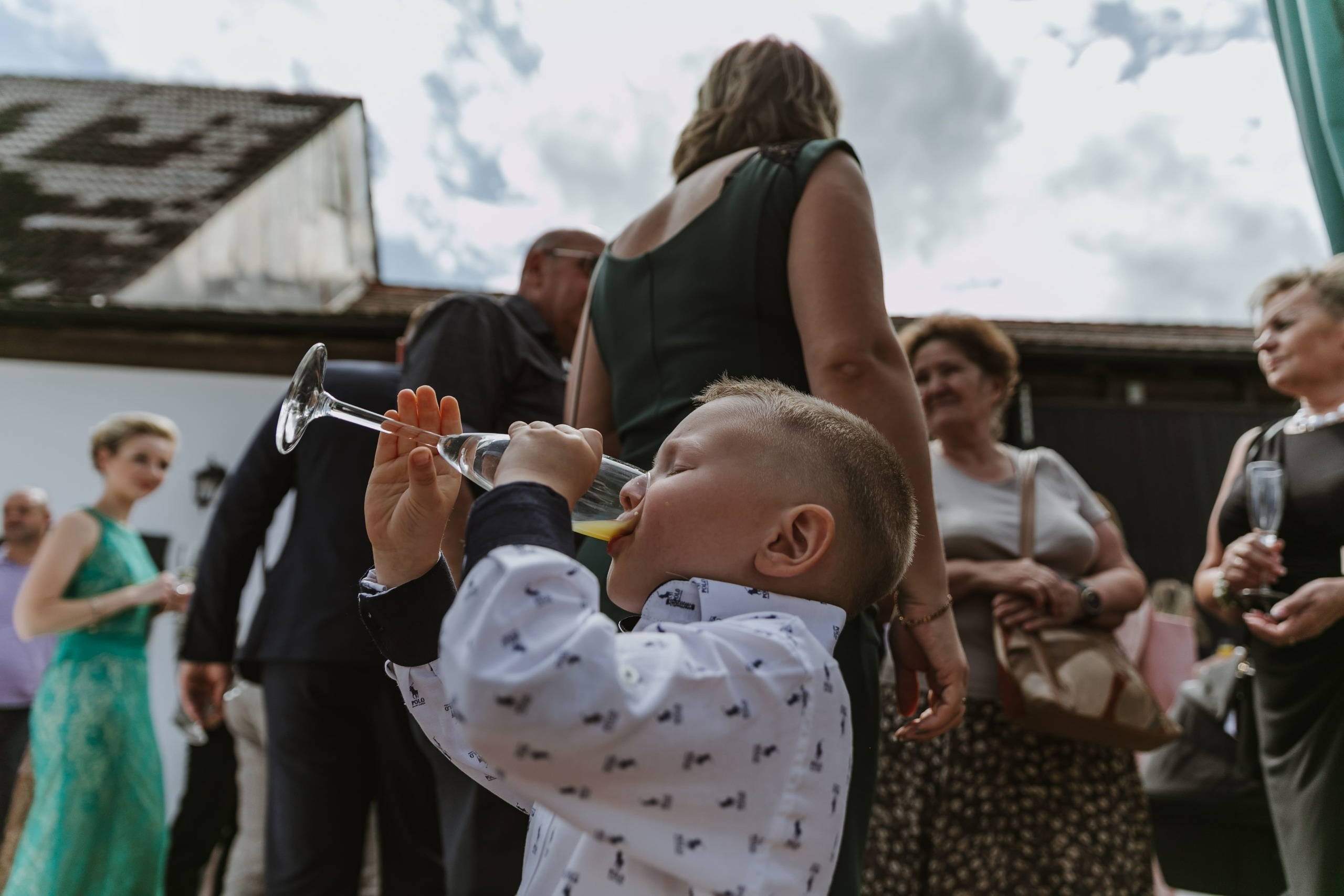 Kleiner Junge trinkt Saft aus einem Champagnerglas vor den Hochzeitsgästen.