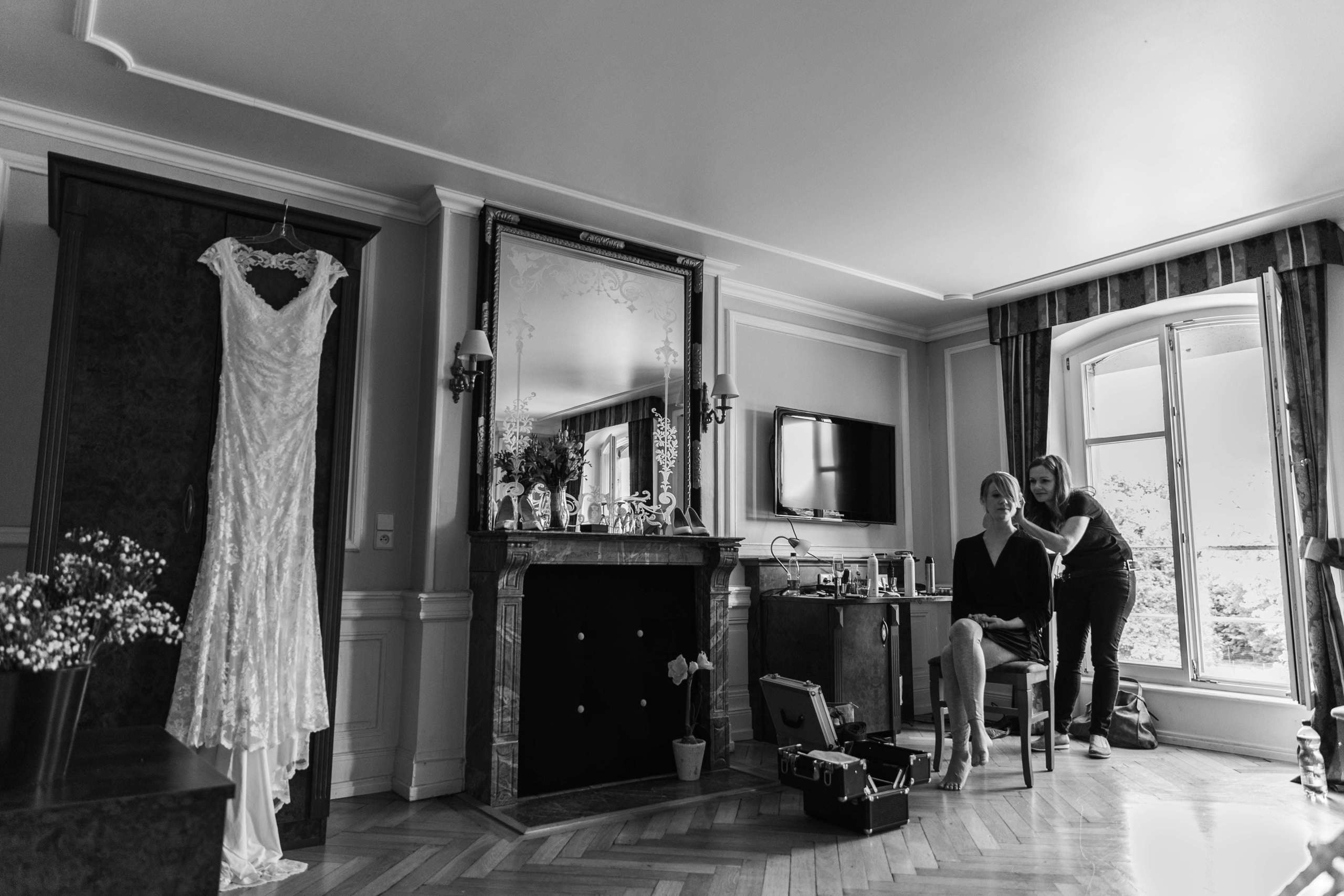 Braut bekommt Frisur, während das Hochzeitskleid neben dem Kamin hängt.