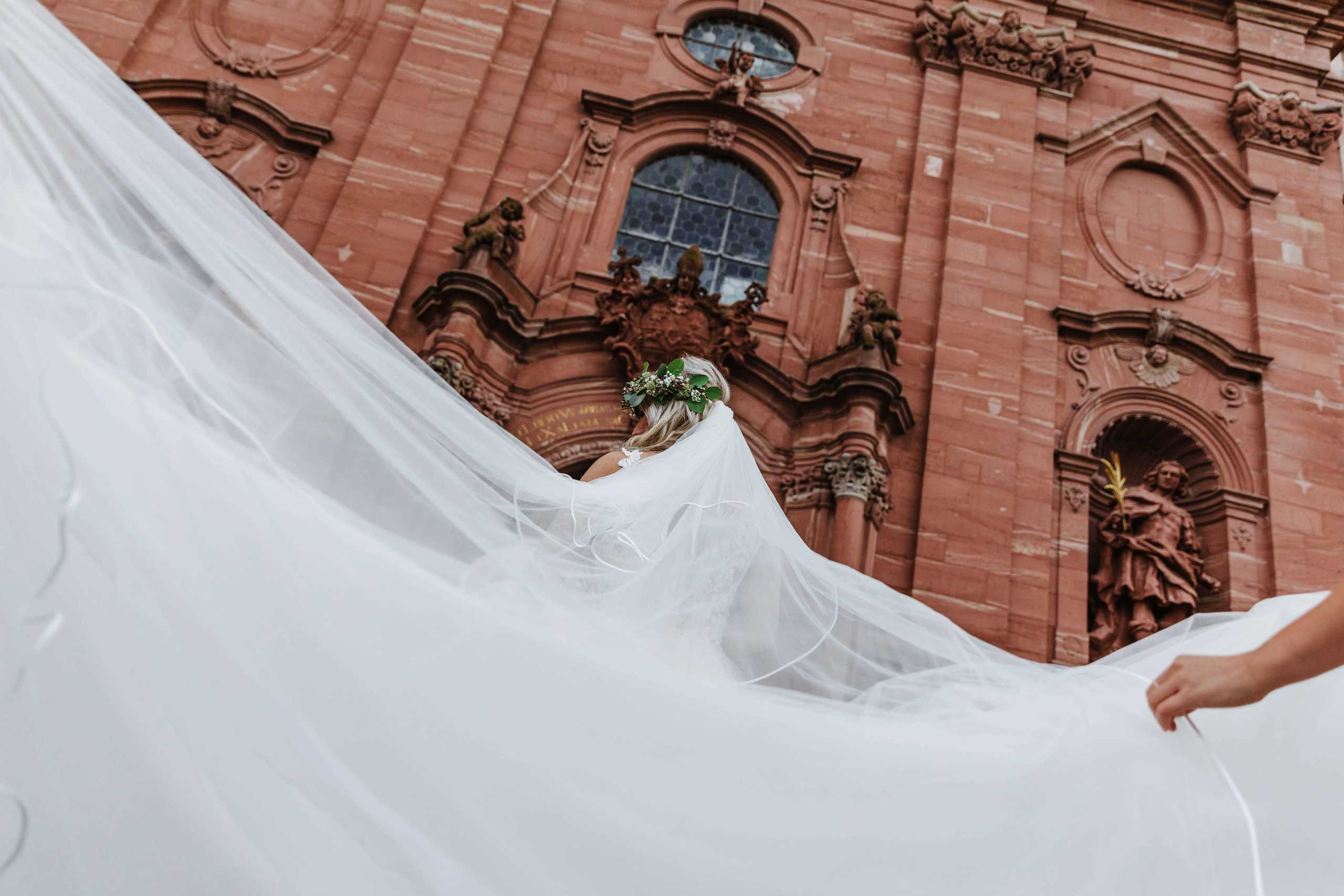 Die Braut geht zur Abteikirche Amorbach, während ihre Freundinnen den atemberaubenden Schleier zurechtrücken.