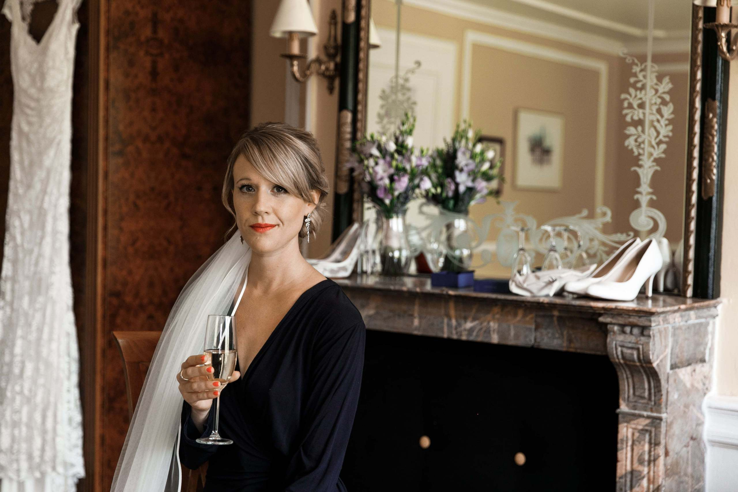 Braut sitzt mit einem Glas Champagner vor ihrem Brautkleid, Brautstrauß und Schuhen.
