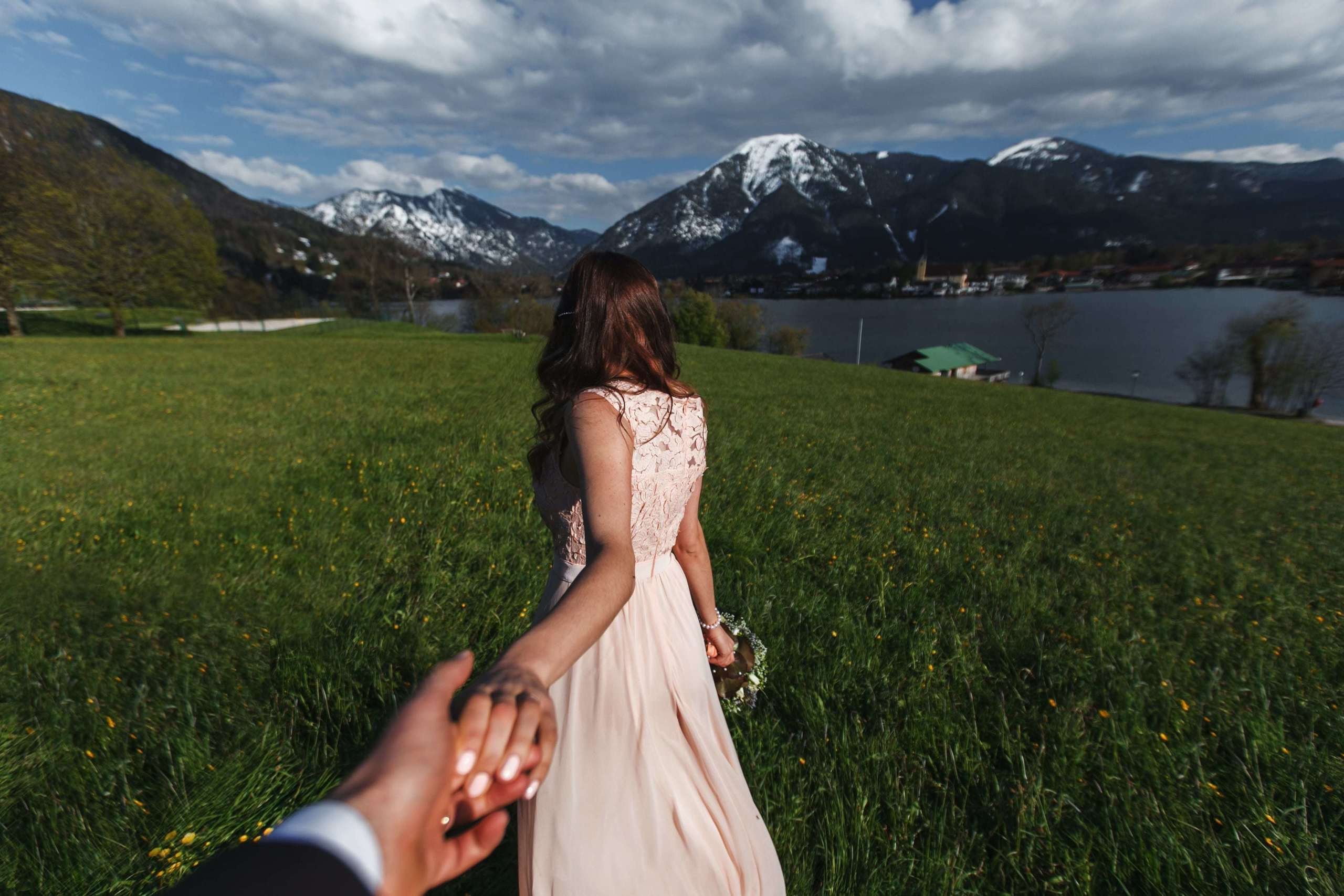 Die Braut führt den Bräutigam an der Hand über eine grüne Wiese zum Tegernsee.