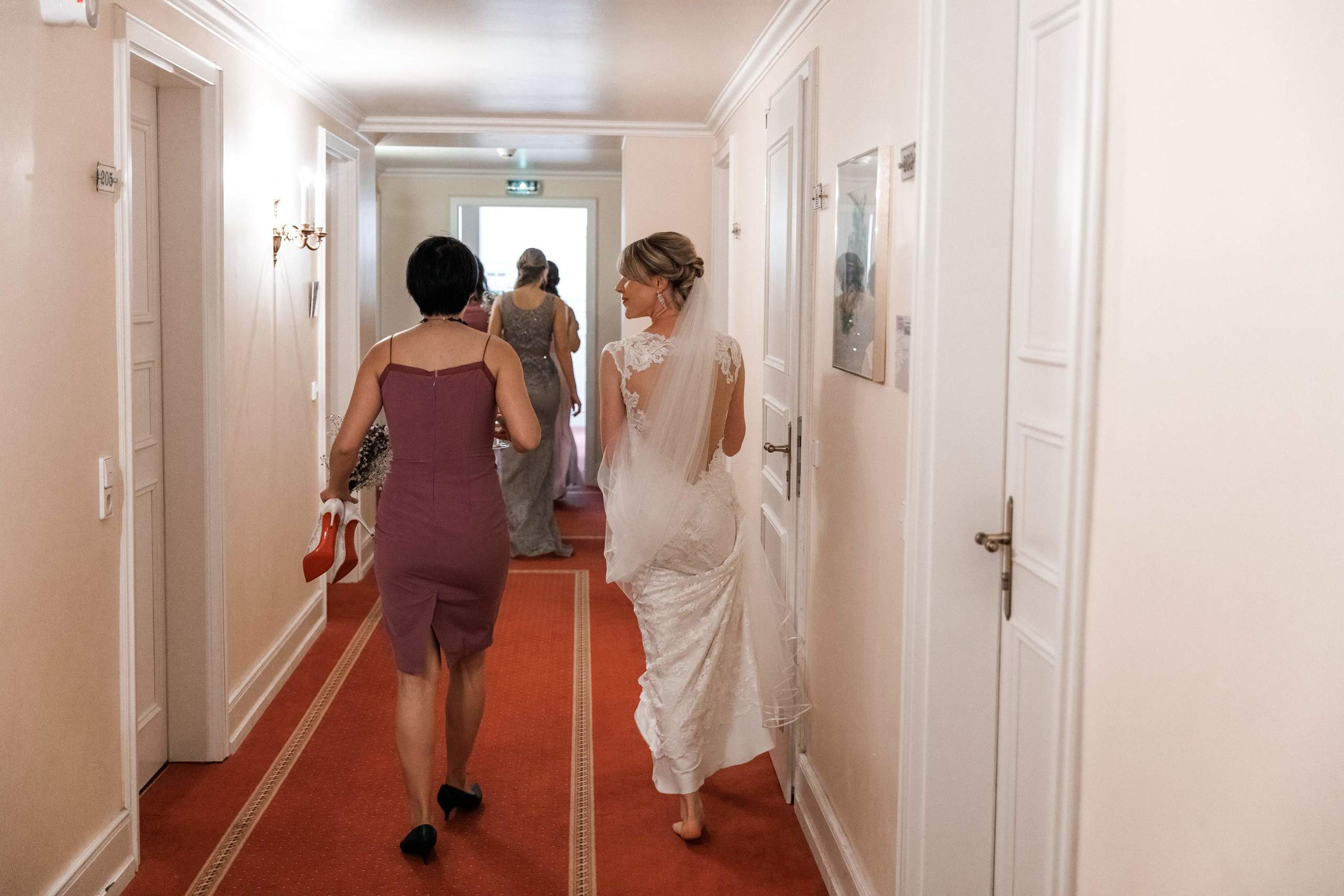 Die Braut geht barfuß den Hotelgang entlang, während ihre Freundin ihre Hochzeitschuhe in der Hand trägt.