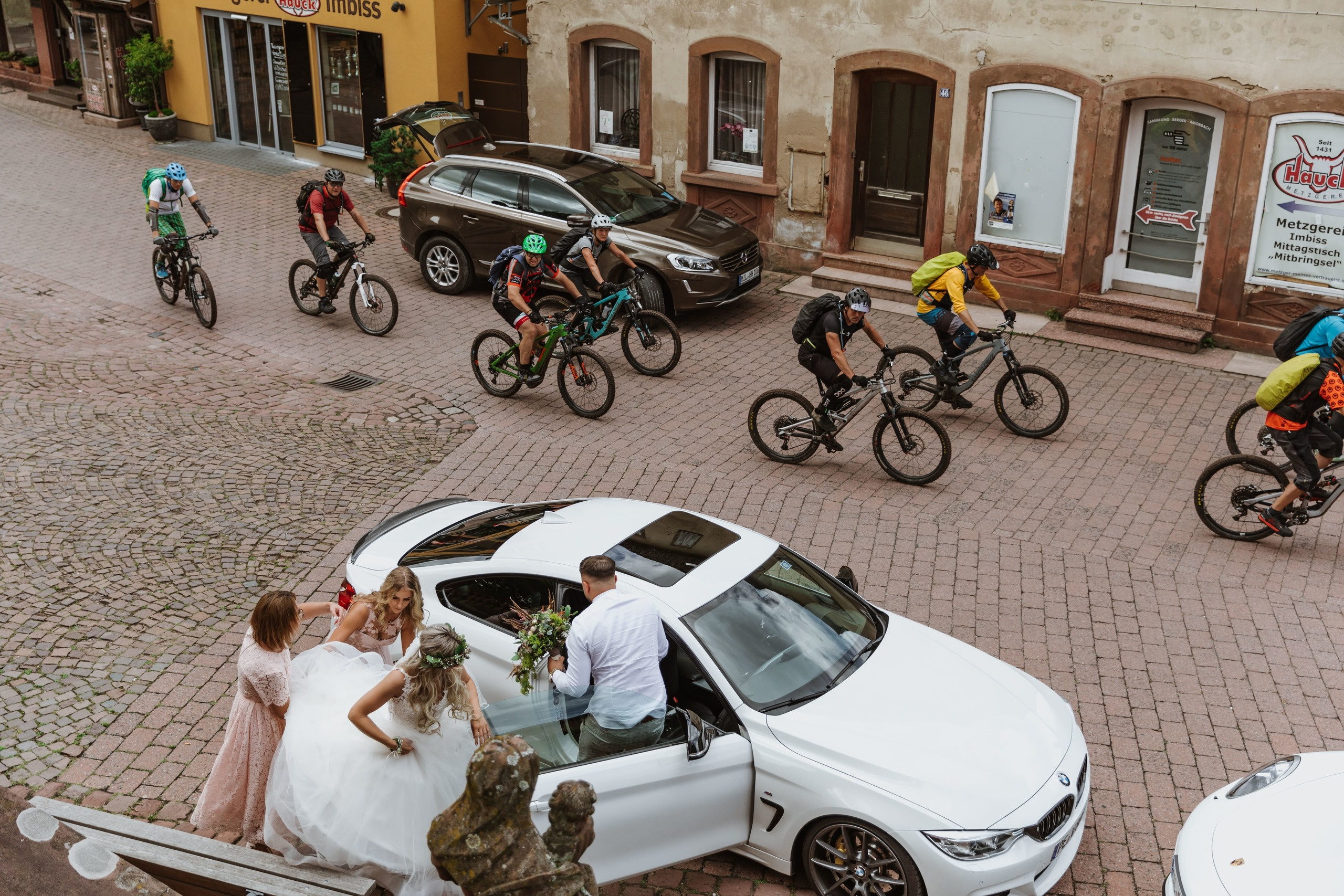 Die Braut steigt vor der Kirche aus dem Auto, während viele Radfahrer vorbeifahren.