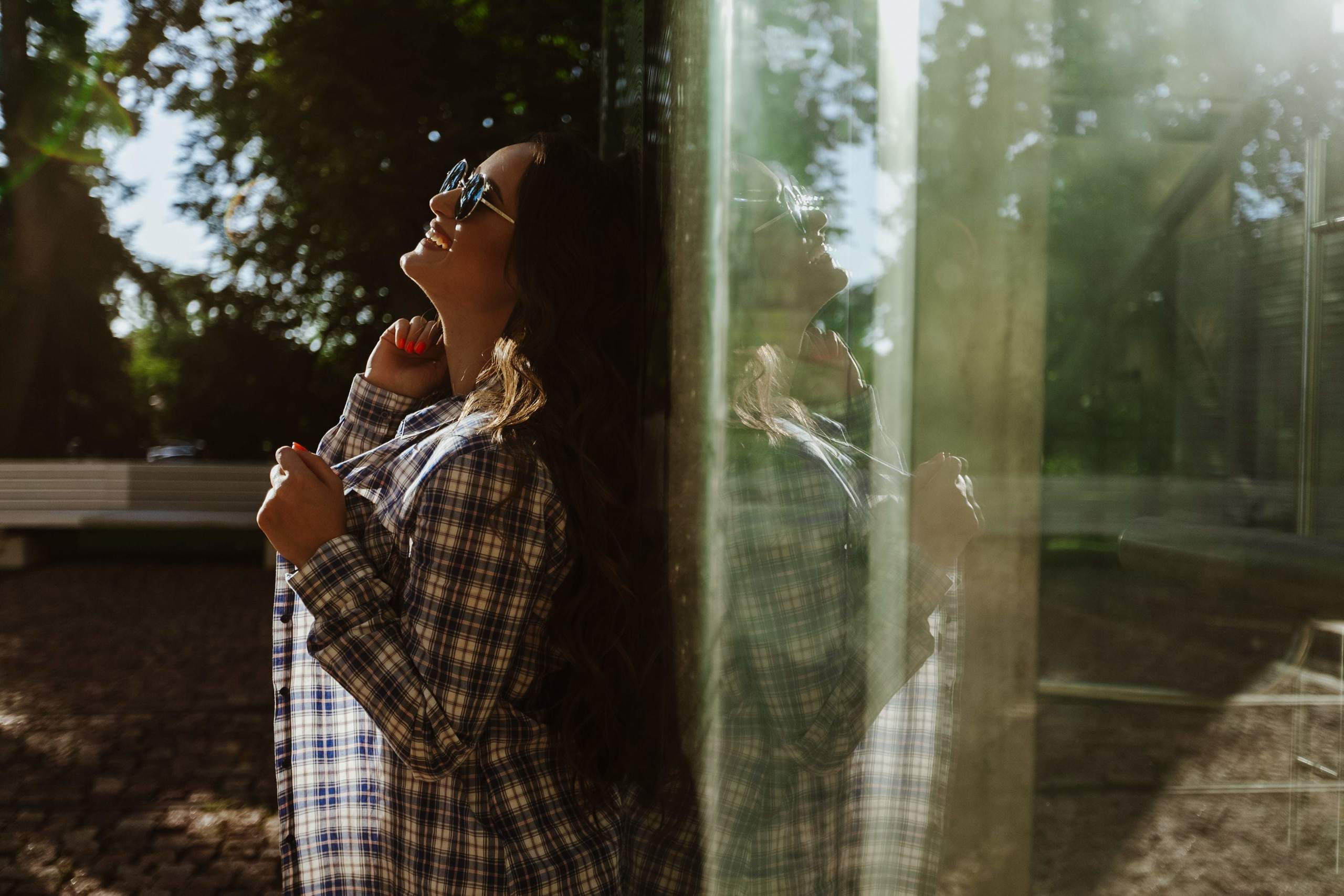 Junges Mädchen steht bei einem Outdoor-Fotoshooting an einer Glaswand, umgeben von grünem Laub und natürlichem Licht.