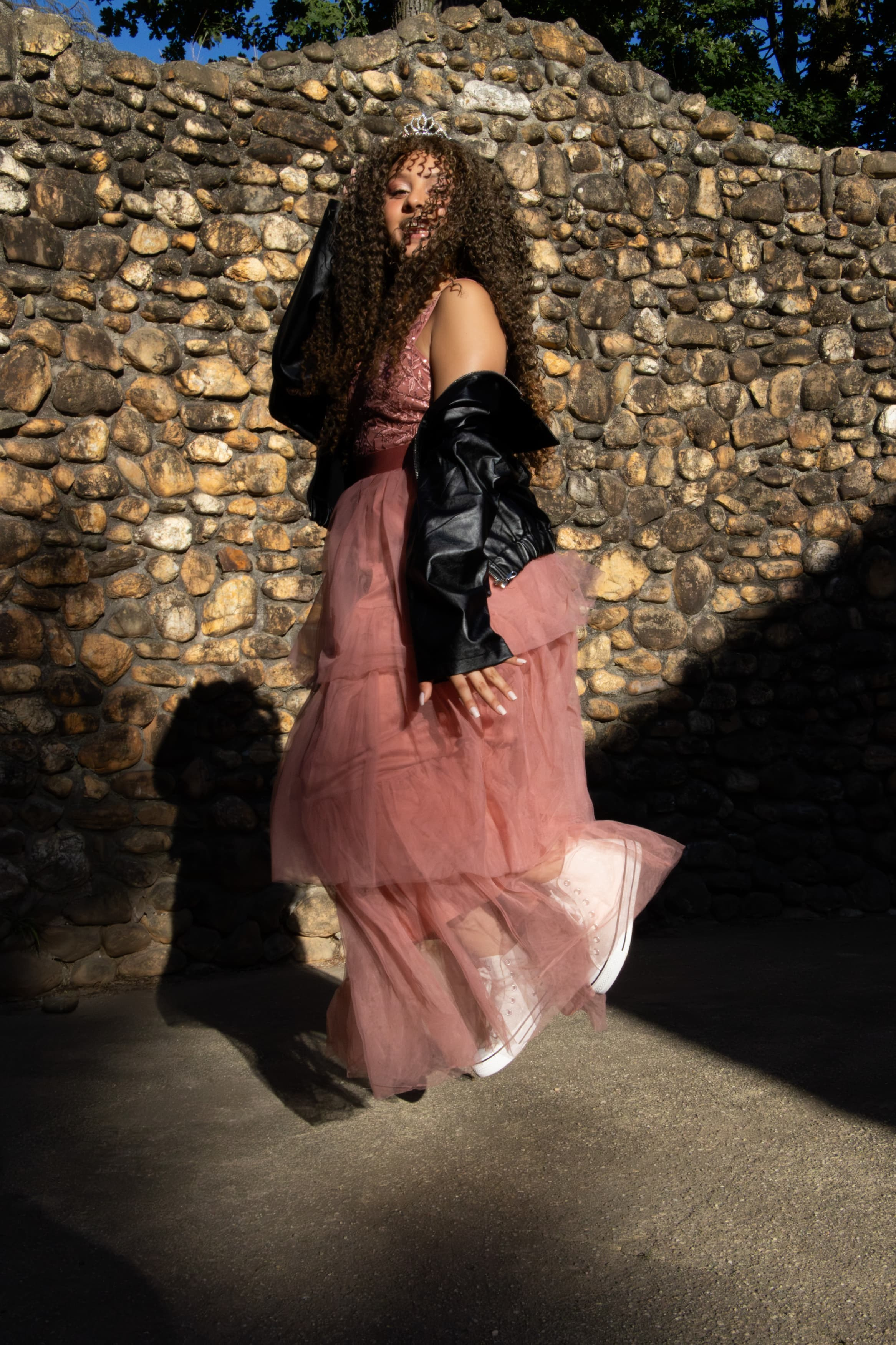 Uma jovem posando em pé ao lado de um muro alto de pedras rústicas, vestindo jaqueta escura e saia rosa longa esvoaçante.