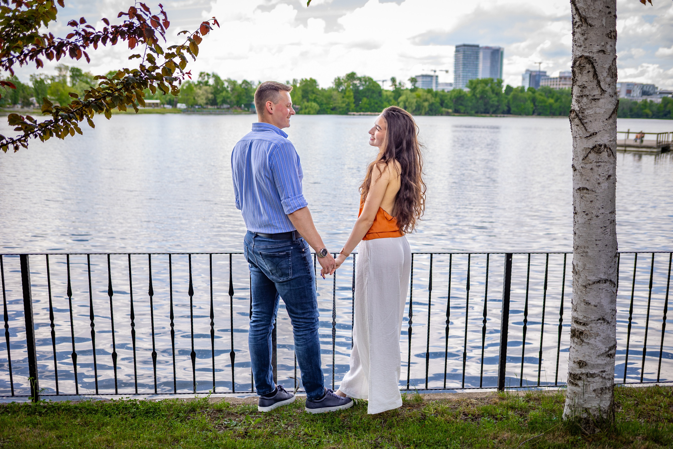Doi tineri se țin de mână și se privesc zâmbind pe malul Lacului Floreasca, într-un portret romantic în aer liber.