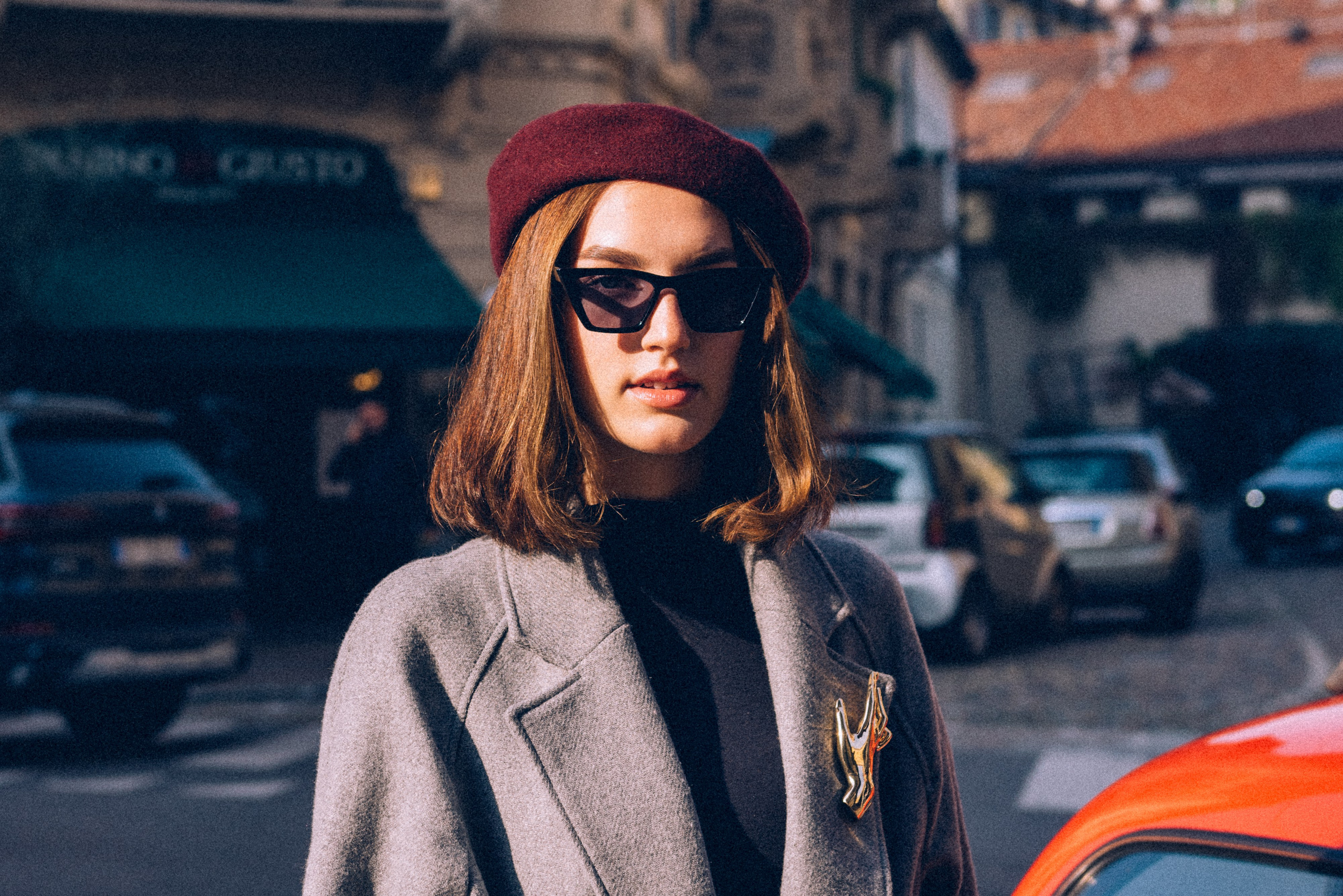 Portrait of a stylish woman in Milan wearing a burgundy beret, statement sunglasses, and a gray coat with a vintage gold brooch, captured under warm urban light