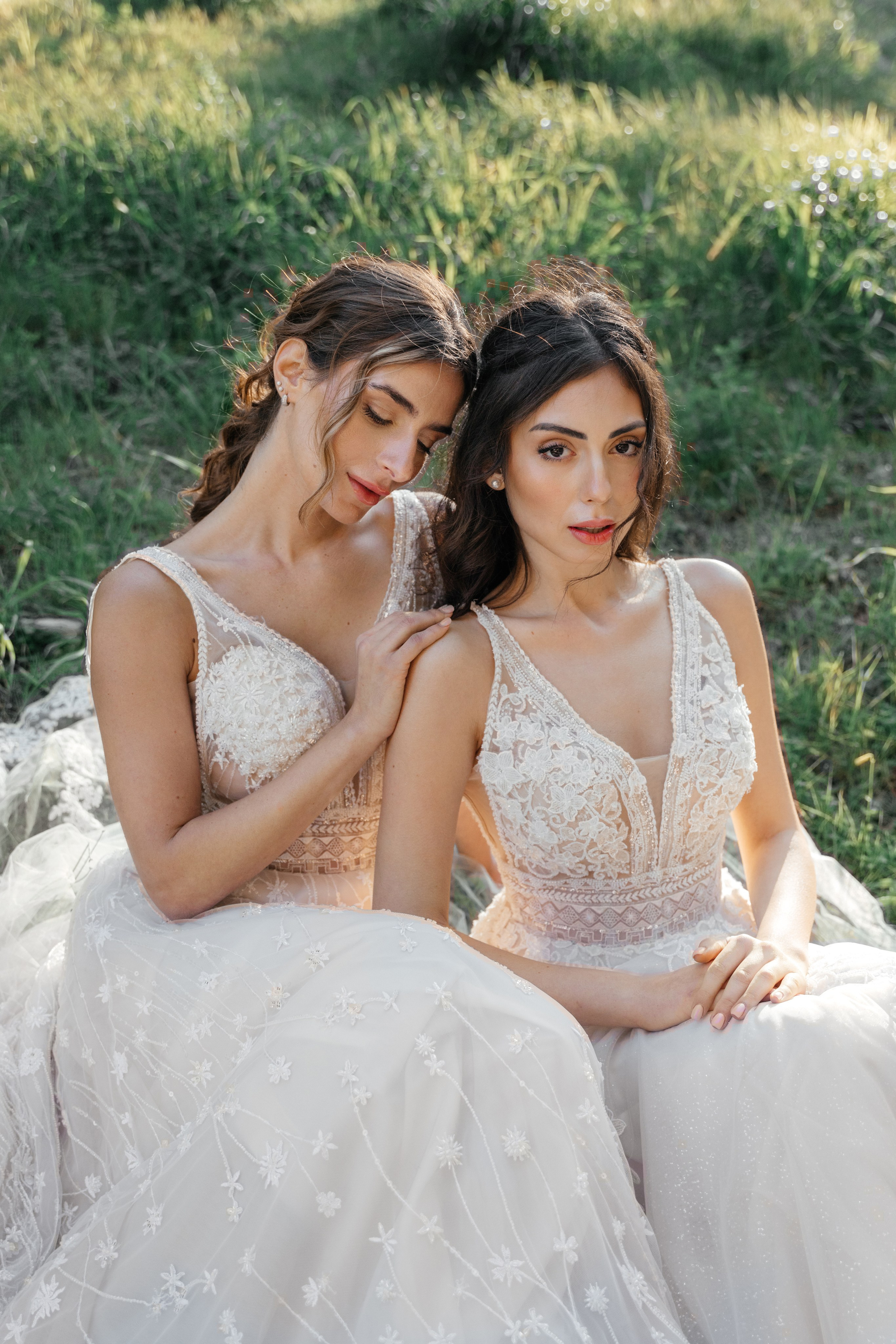 Two brides in elegant wedding dresses pose in the narrow, cobblestone streets of Rhodes' Old Town, surrounded by historic stone buildings and vibrant bougainvillea. The editorial-style photo captures their intimate connection and the timeless charm of the medieval setting, with soft natural light enhancing the romantic atmosphere.