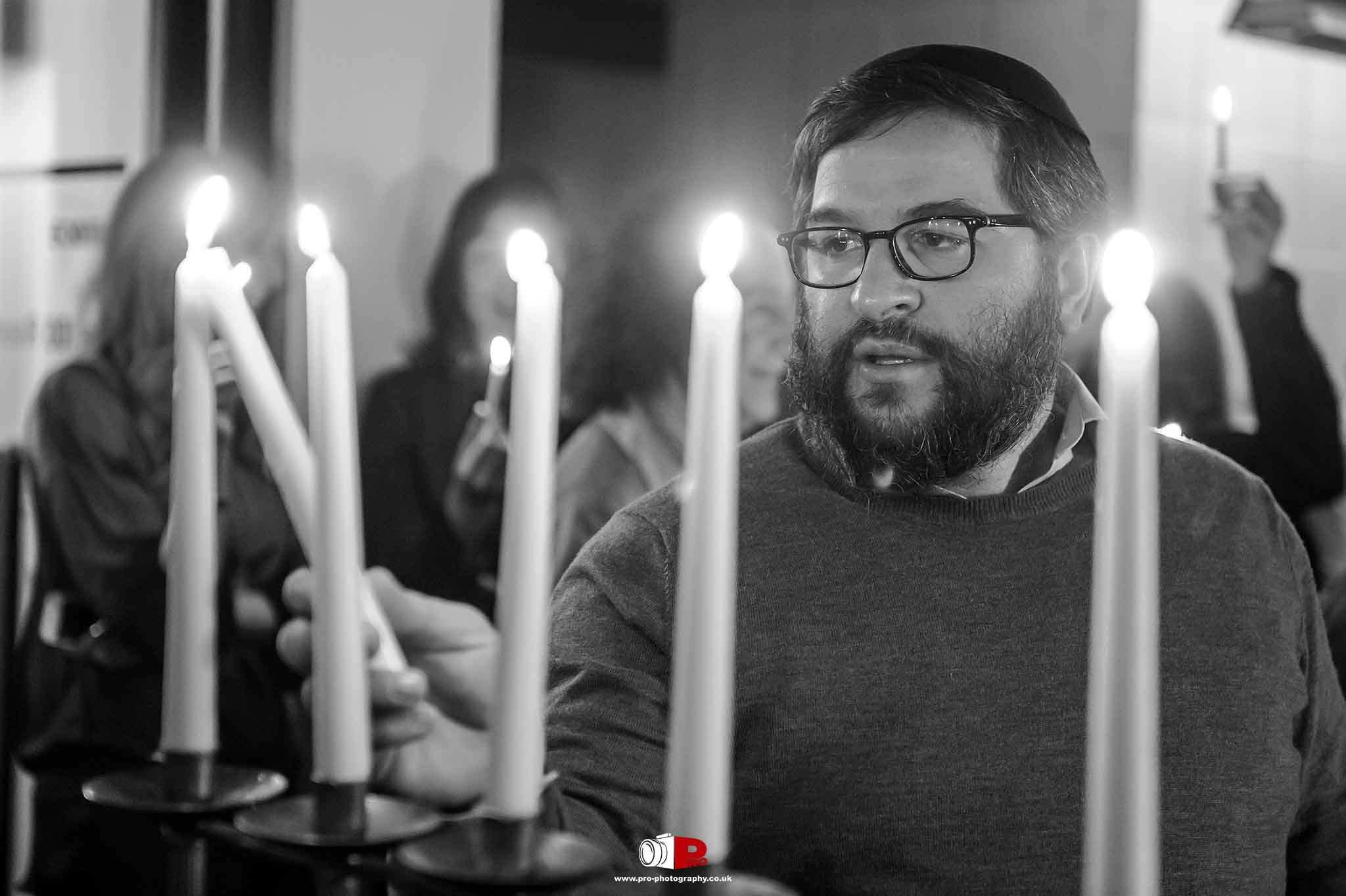 A man is lighting candles during a Hanukkah ceremony, surrounded by attendees holding candles.