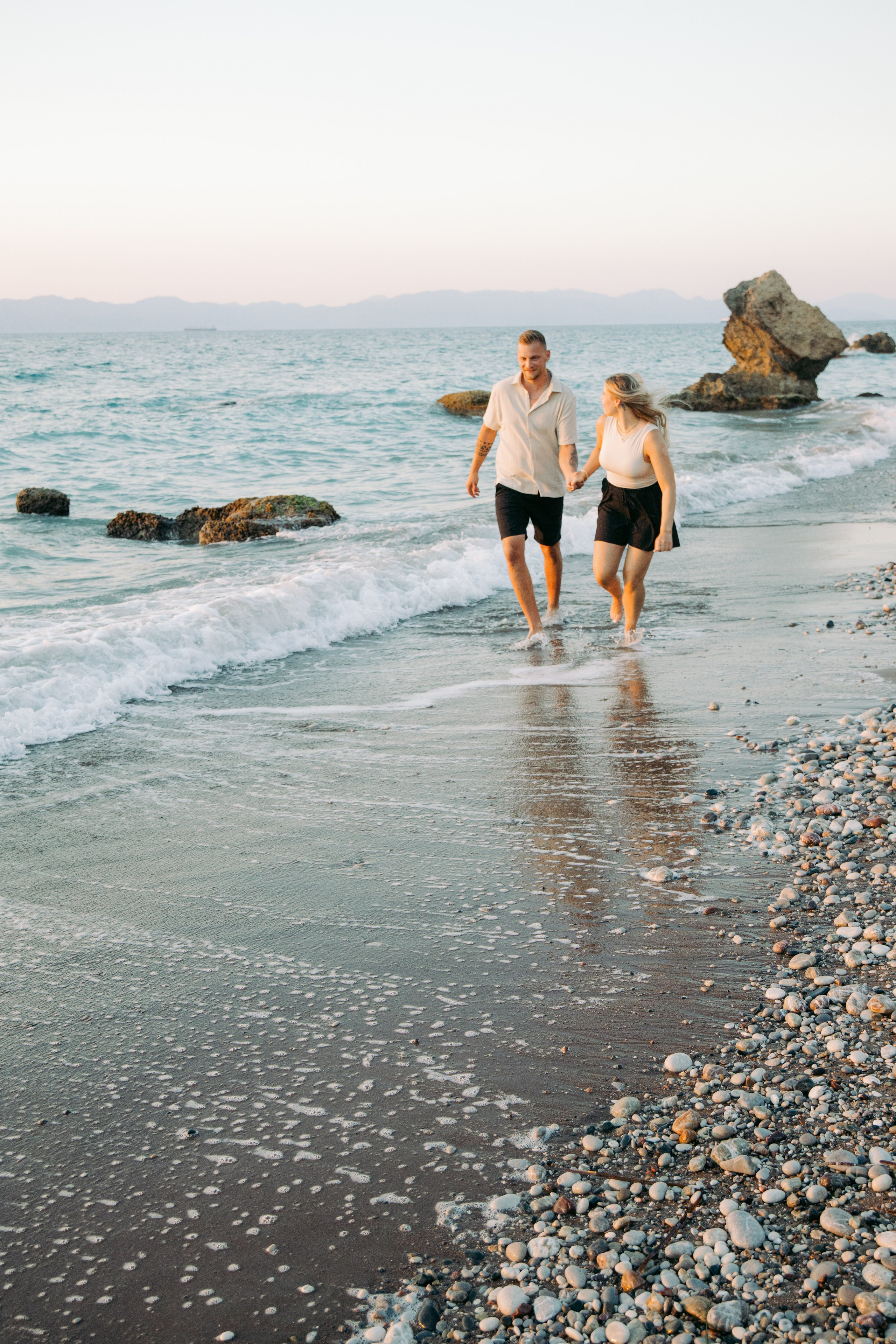 "Ein Paar spaziert Hand in Hand am steinigen Ufer von Kato Petres auf Rhodos entlang. Sie gehen direkt am Wasser, während im Hintergrund die hohen, zerklüfteten Kalksteinfelsen aufragen und das blaue Meer sanft gegen die Kiesel brandet."