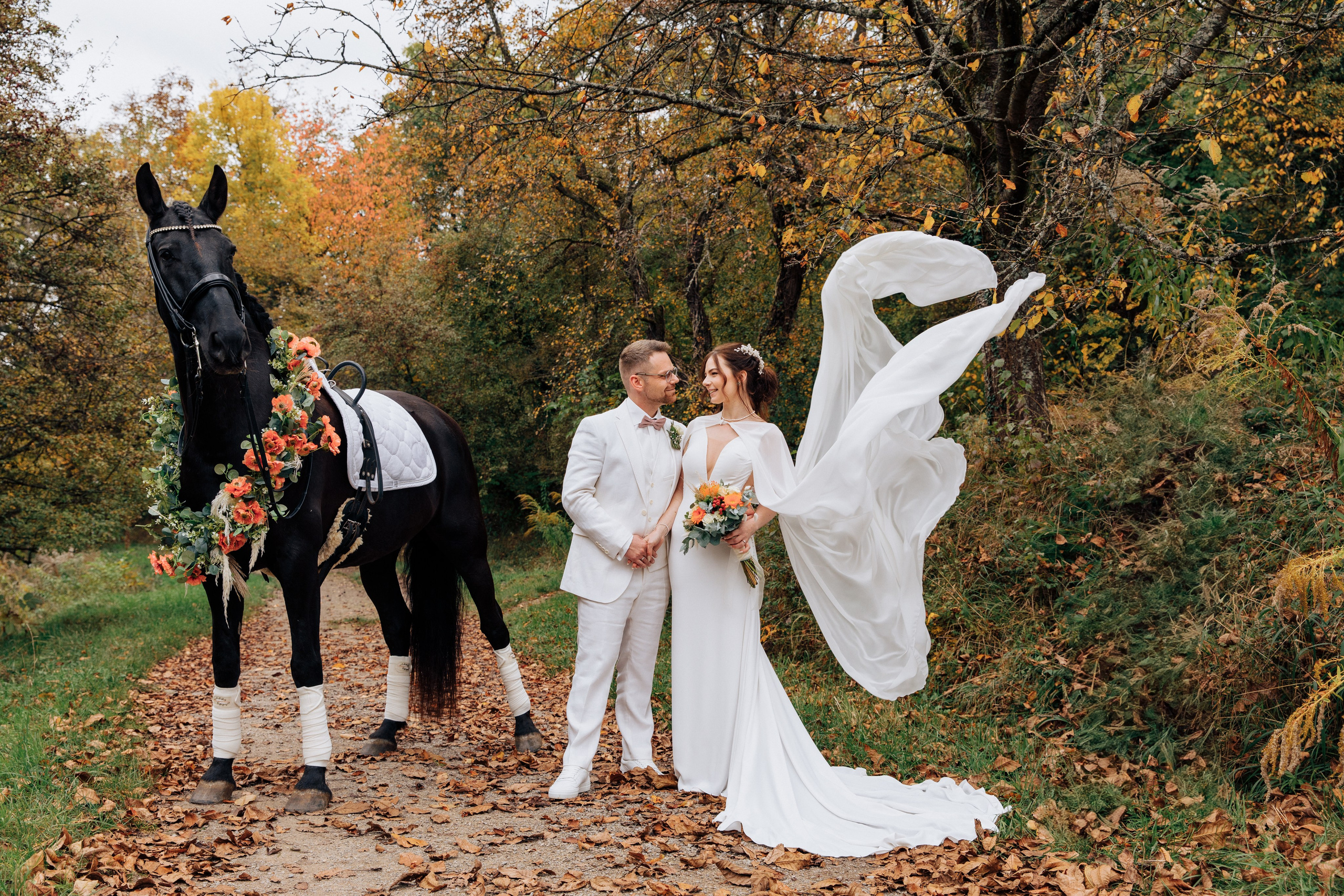 Das Brautpaar schaut sich verliebt an, neben ihnen steht ein Pferd, und das Hochzeitskleid der Braut weht romantisch im Wind. Perfekte Hochzeitsfotos.