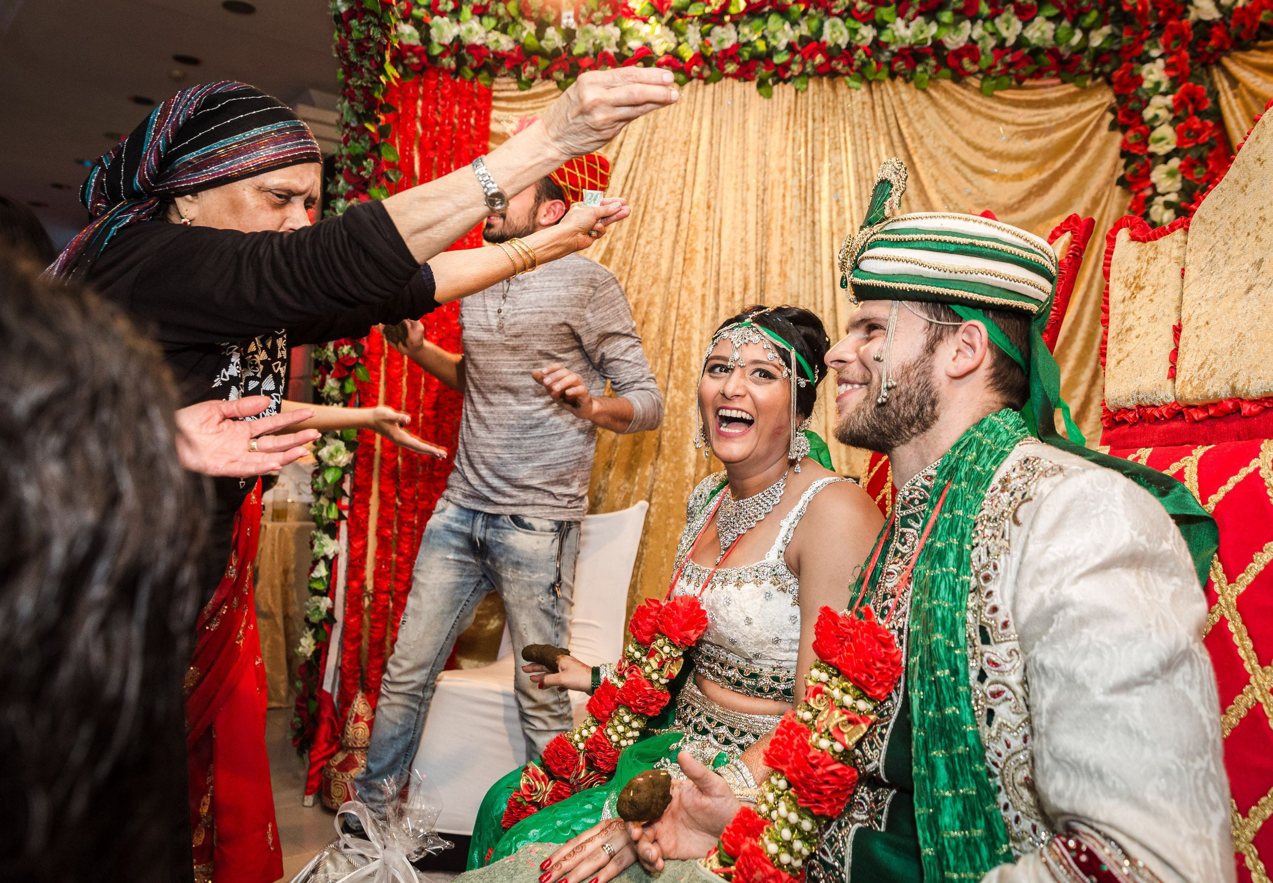A vibrant photo of an Indian wedding ceremony, showing a woman throwing rice over the bride and groom as part of a traditional blessing. The couple, dressed in colorful wedding attire, are seated under a decorated canopy, surrounded by bright floral arrangements and festive decorations. The moment captures the joy and cultural significance of the celebration.
