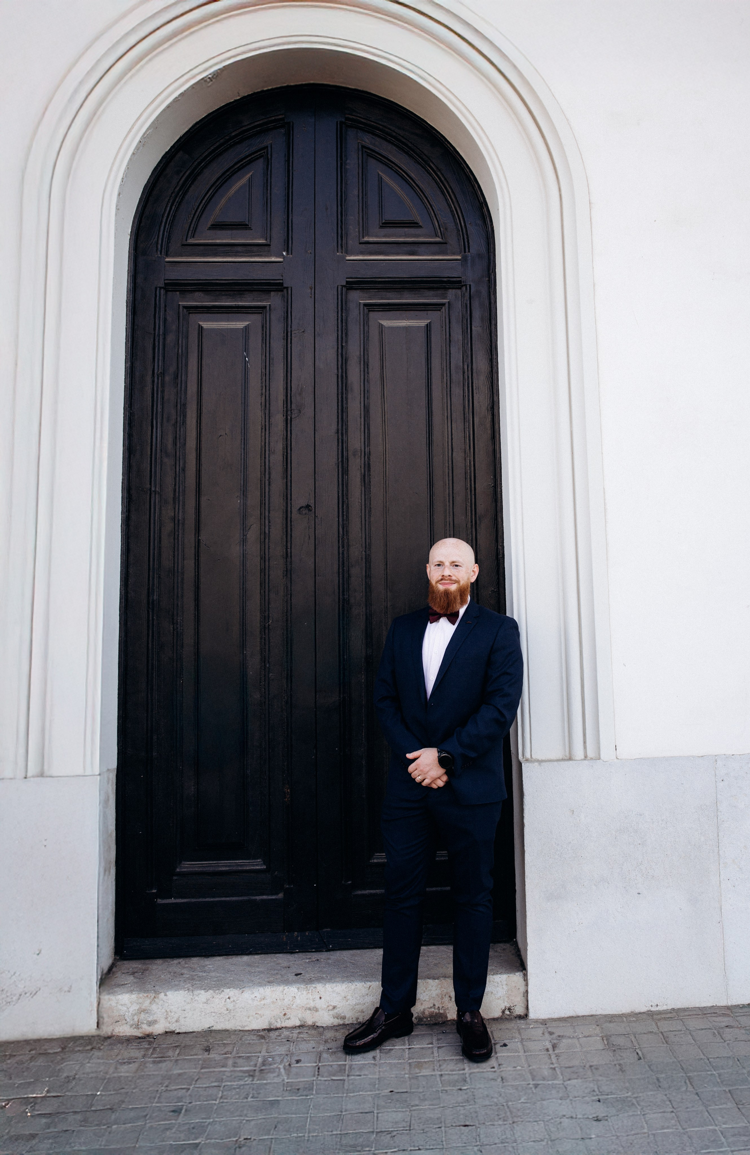 Groom in a dark suit stands confidently in front of a grand arched doorway in the historic center of València. This elegant civil wedding portrait highlights timeless architecture and modern destination wedding style in Spain.