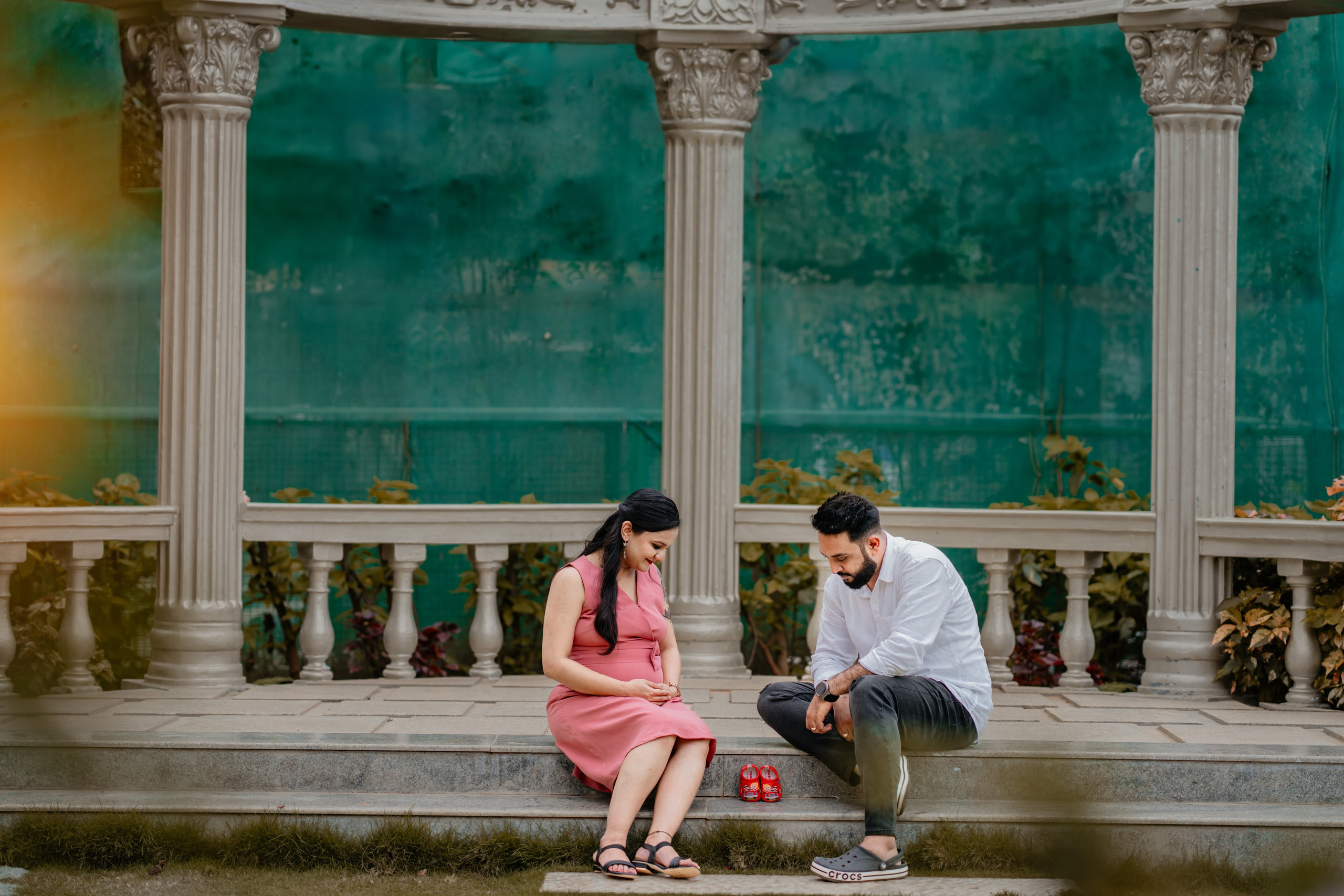 Outdoor maternity photoshoot in Bengaluru featuring a couple sitting in a classical stone gazebo with white pillars and tiny red baby shoes.