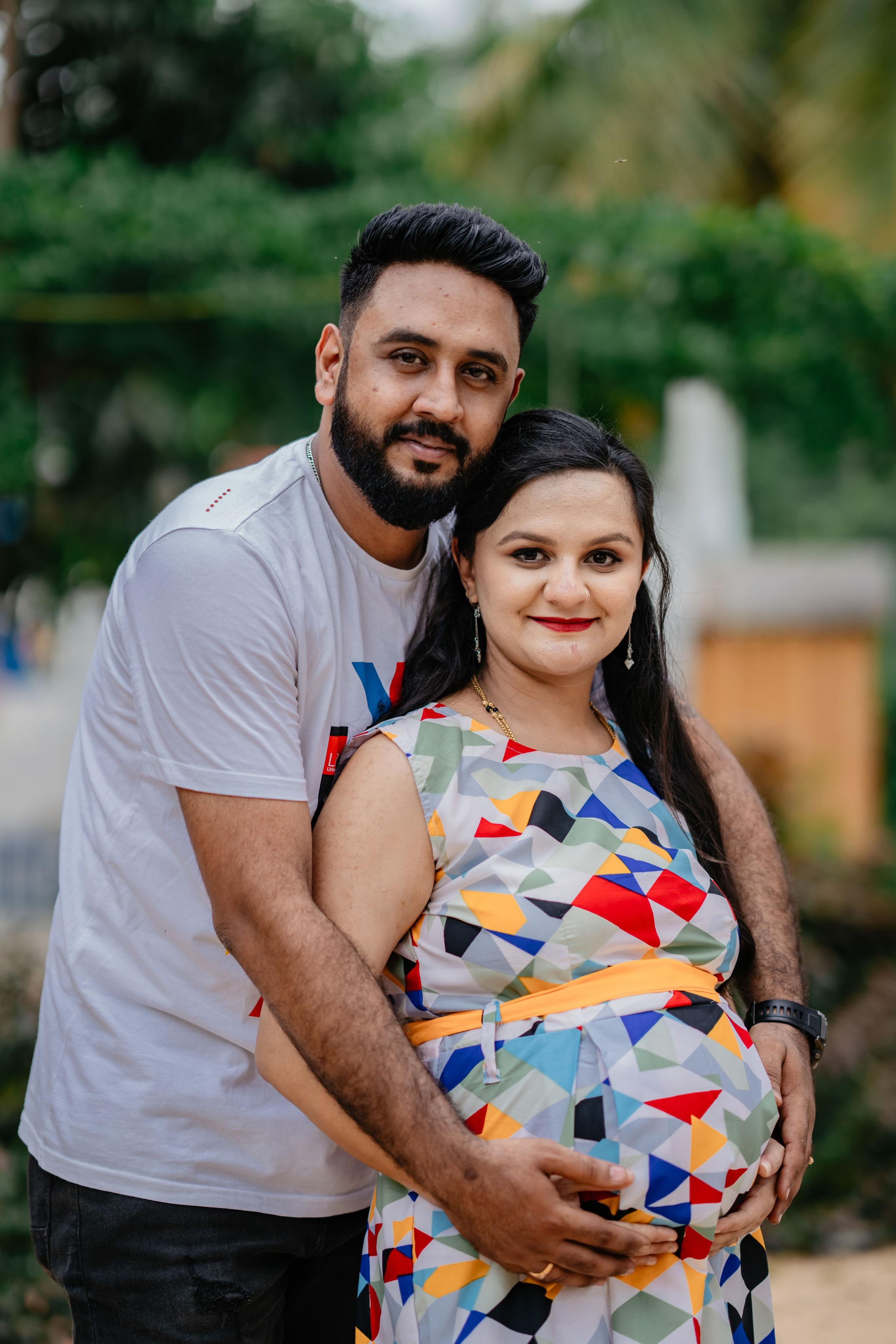 Outdoor maternity photoshoot in Bengaluru featuring a man in a white t-shirt hugging a woman in a colorful geometric print dress from behind.