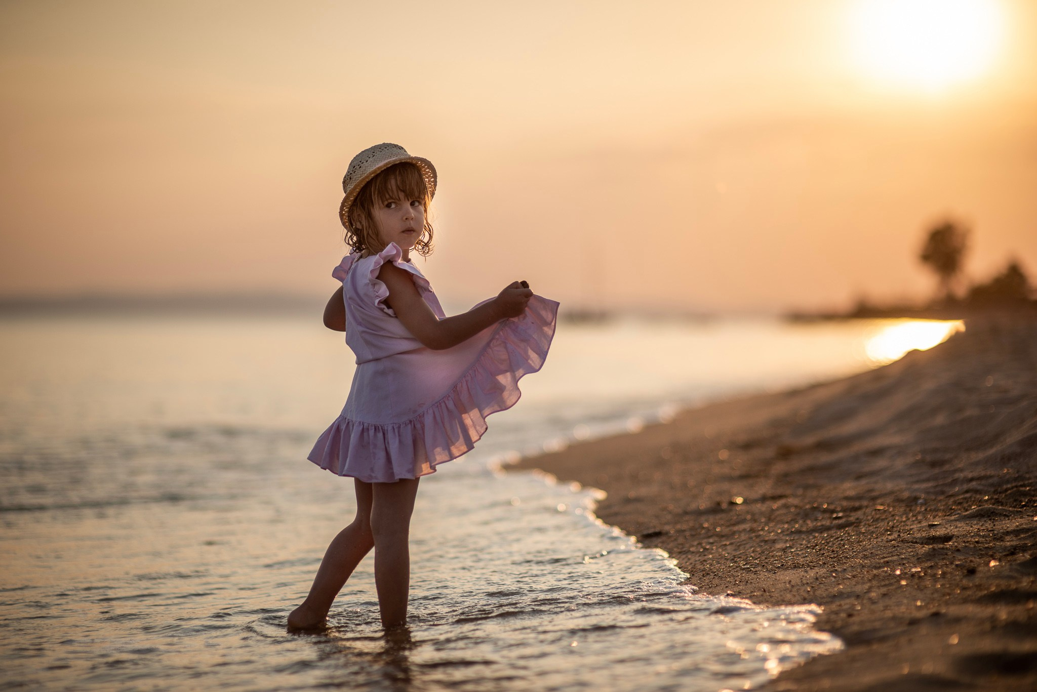 Mlada devojčica u roze haljini i slamnatom šeširu, bosa stoji u plićaku na plaži tokom zalaska sunca u zlatnom satu.