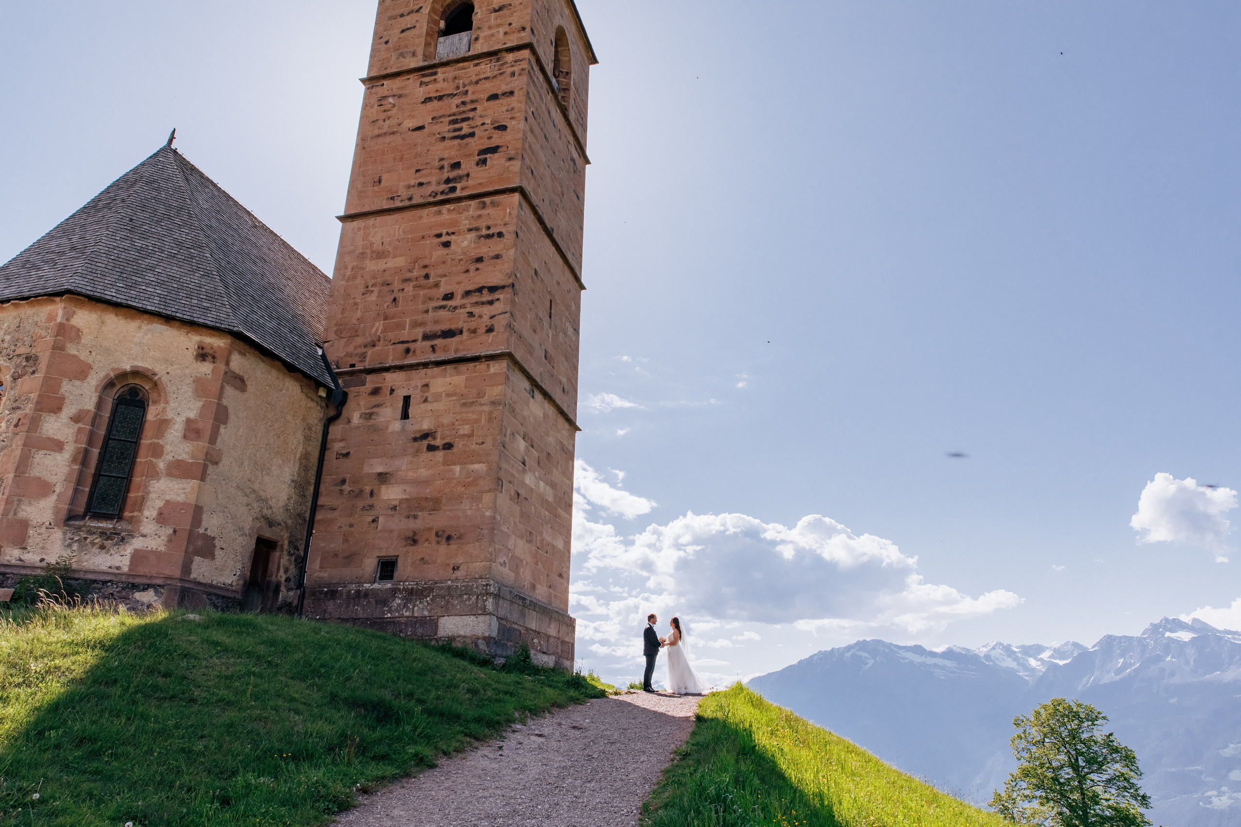 Das Brautpaar steht Hand in Hand neben der St. Kathrein-Kirche in Hafling vor atemberaubender Bergkulisse.