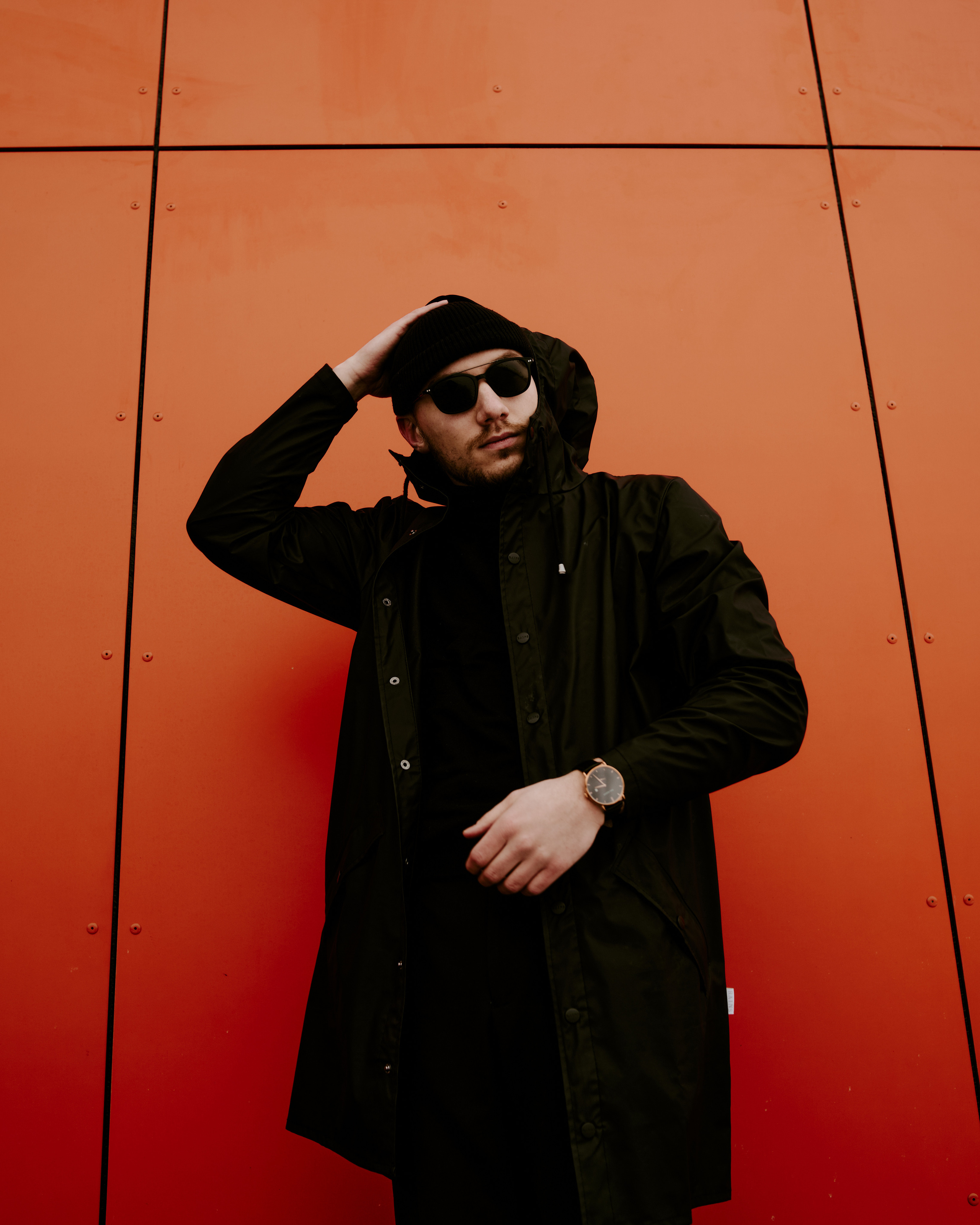 A person posing against a bold orange wall, wearing dark sunglasses and a stylish black outfit, exuding urban confidence.