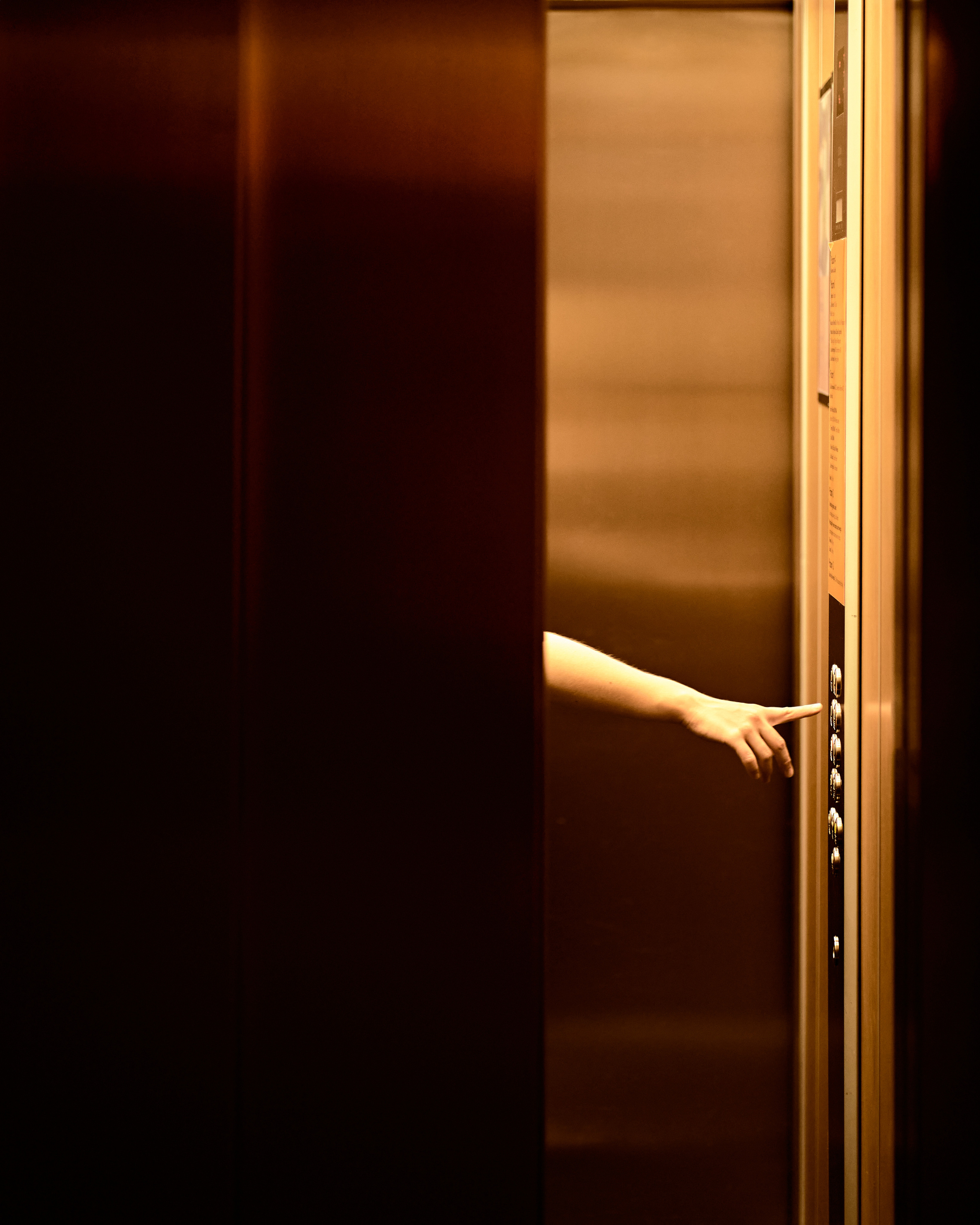 A minimalist shot of a hand emerging from an elevator door, surrounded by warm metallic tones.