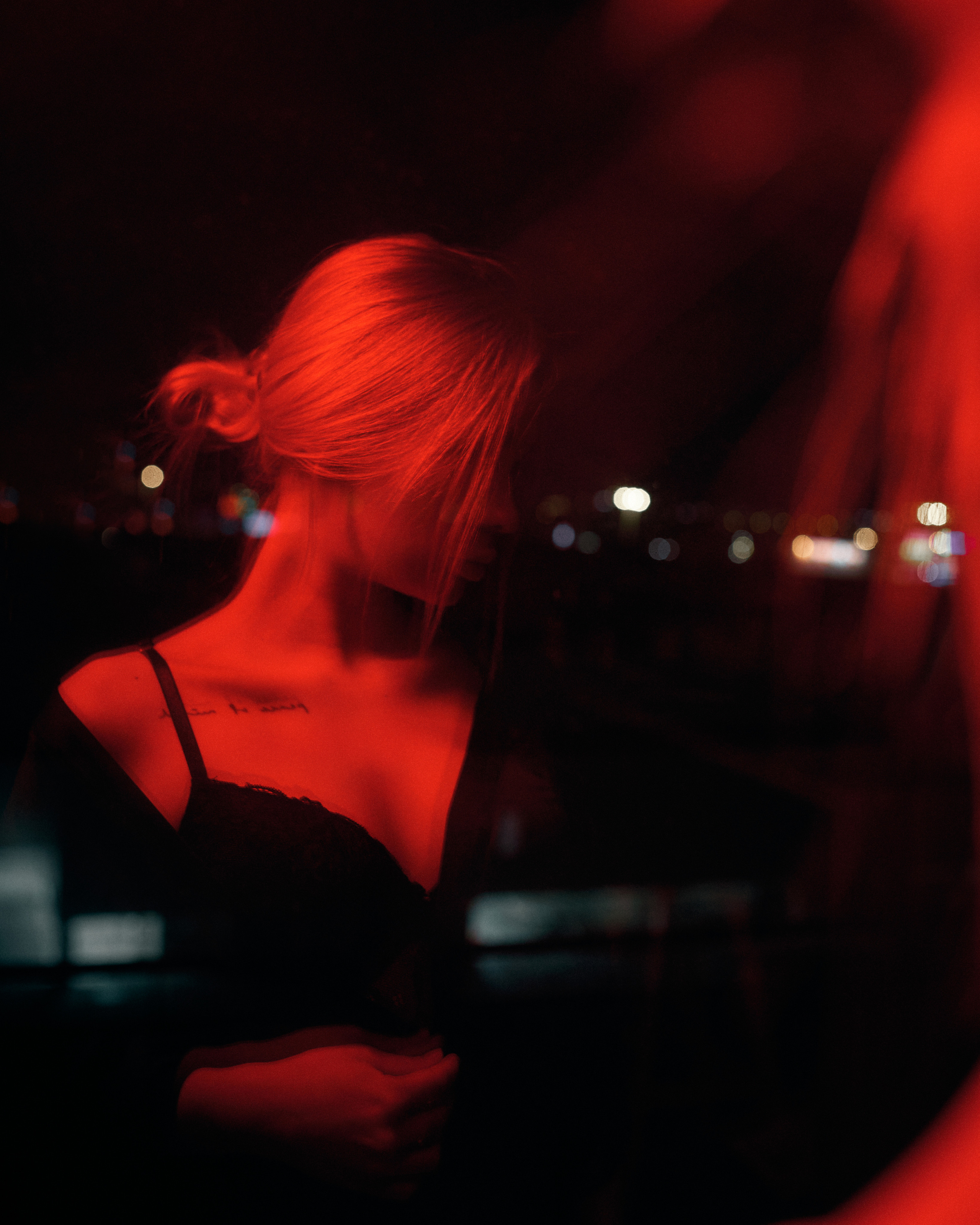 A close-up of a woman in black lingerie, bathed in soft red lighting, with a blurred cityscape behind her, evoking a sensual and mysterious mood.