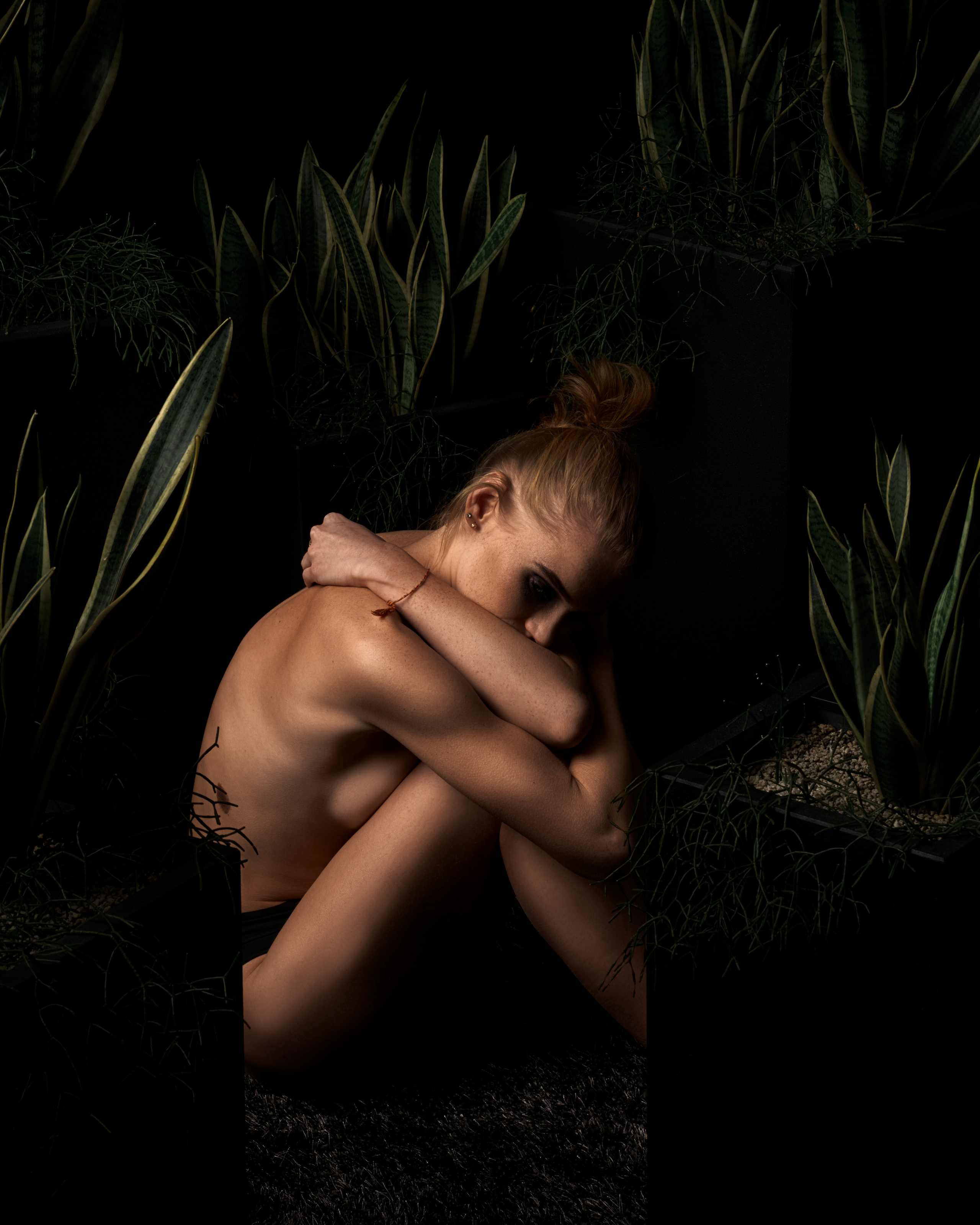 A nude woman curled up among tall plants, highlighted by soft directional lighting in a natural yet intimate setting.