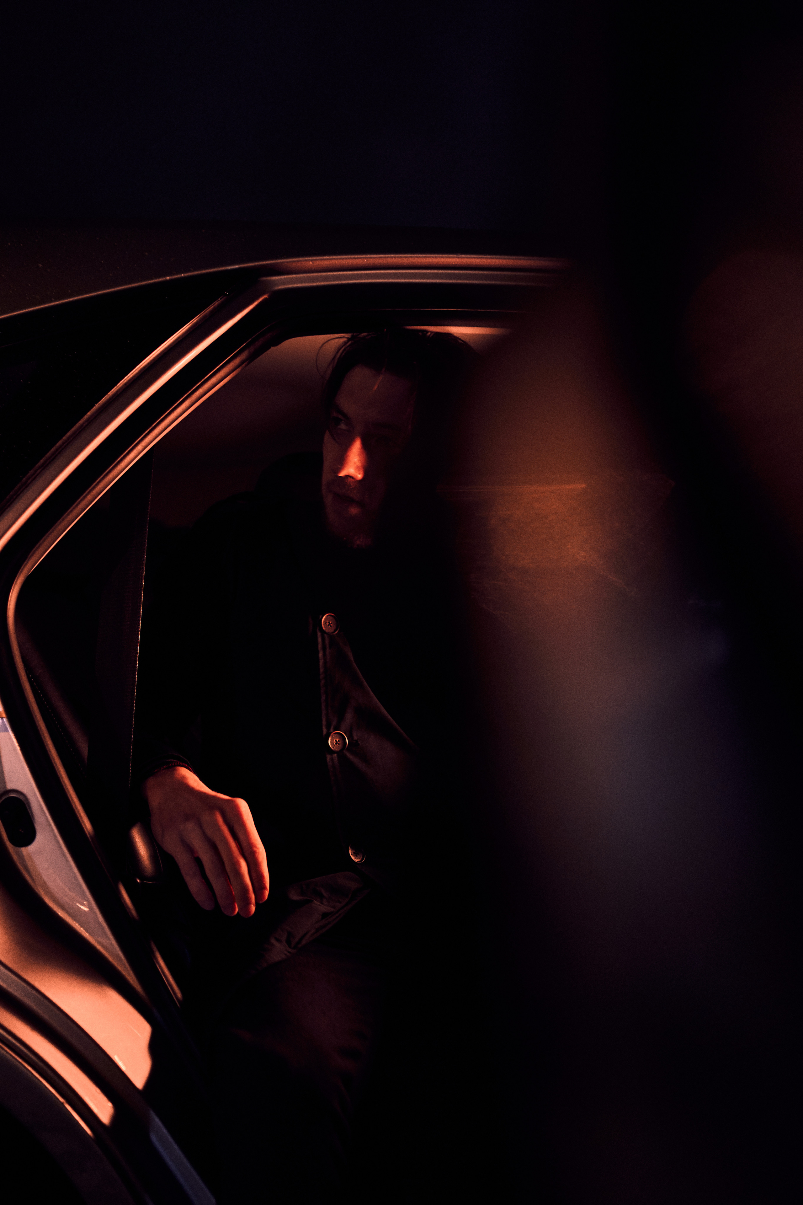 A man seated in a luxury car, partially obscured by shadows and reflections, creating a sophisticated and enigmatic moo