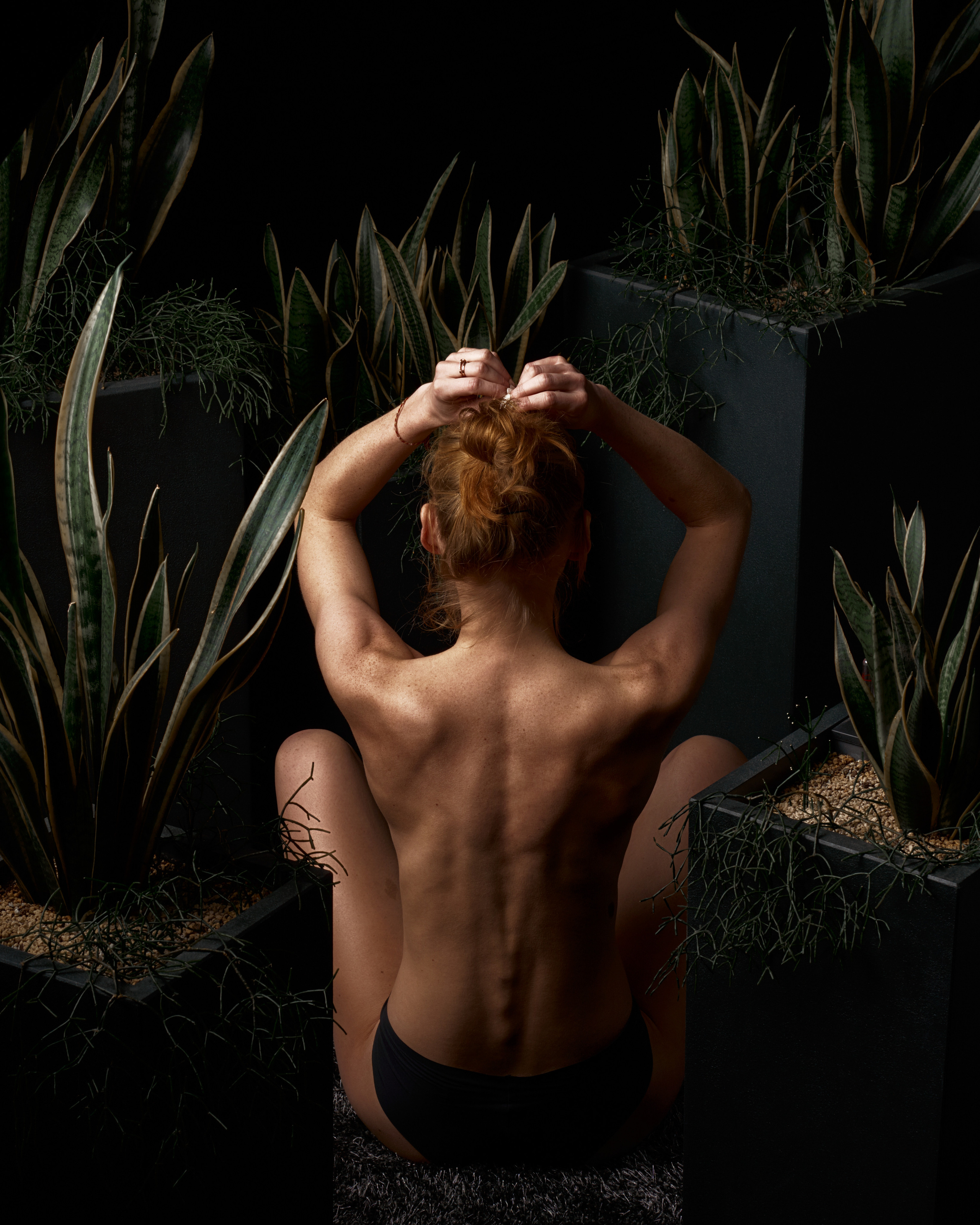 A rear view of a shirtless person sitting among tall plants, with their arms raised to tie their hair, creating a natural and intimate composition.