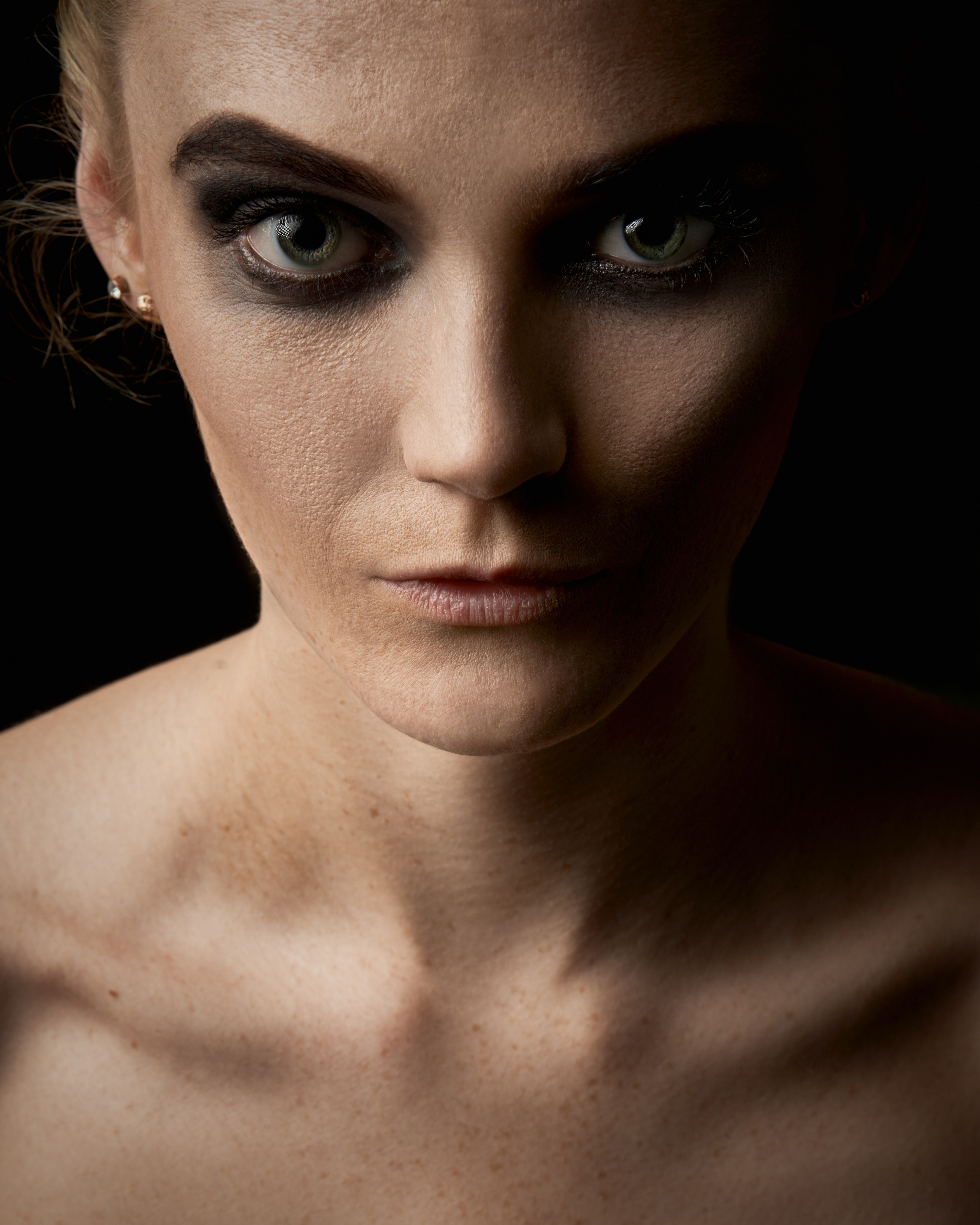 A close-up portrait of a woman with bold makeup, her intense gaze emphasized by soft lighting and a dark background.