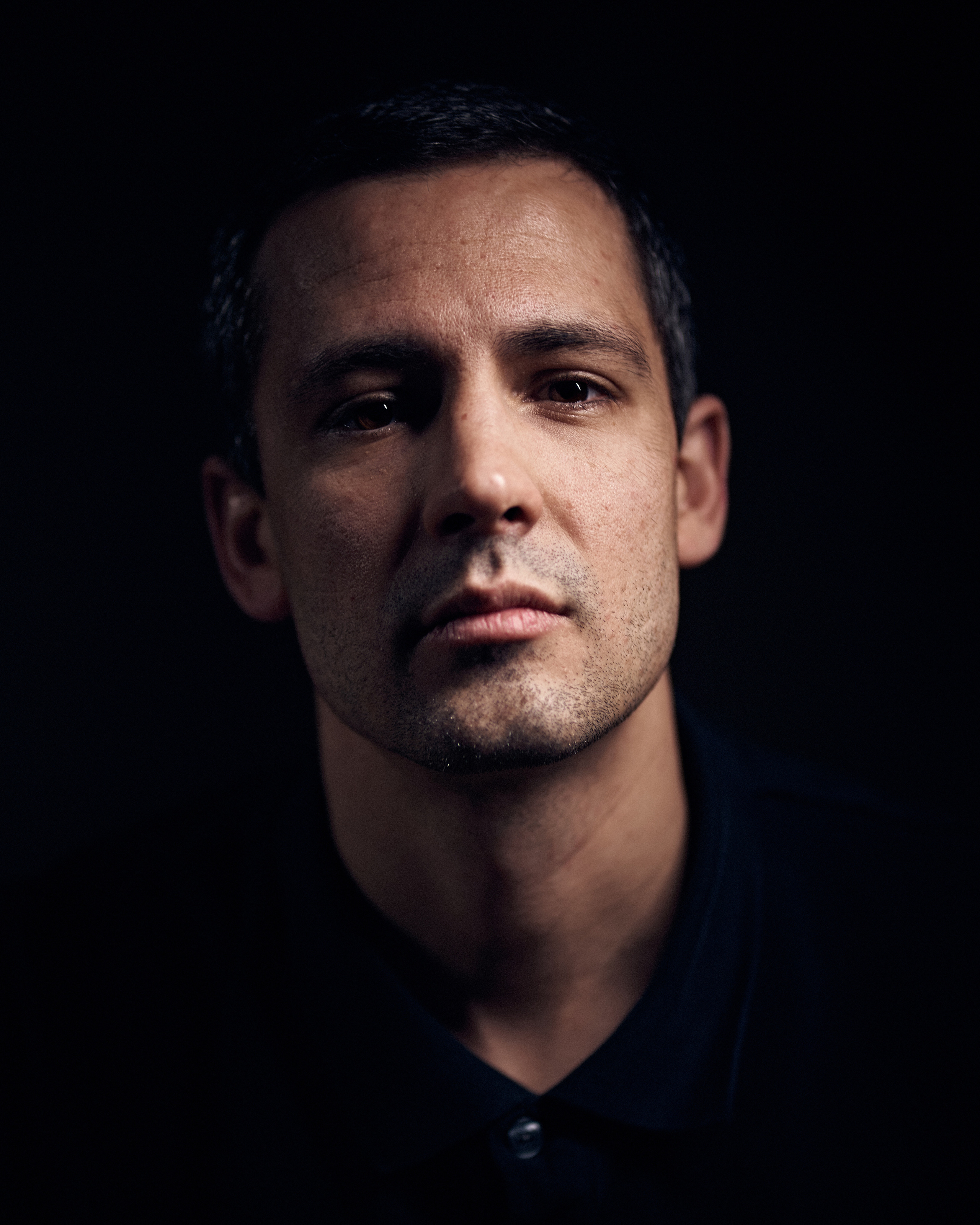 A studio portrait of a man with a serious expression, captured under soft directional lighting against a dark background.