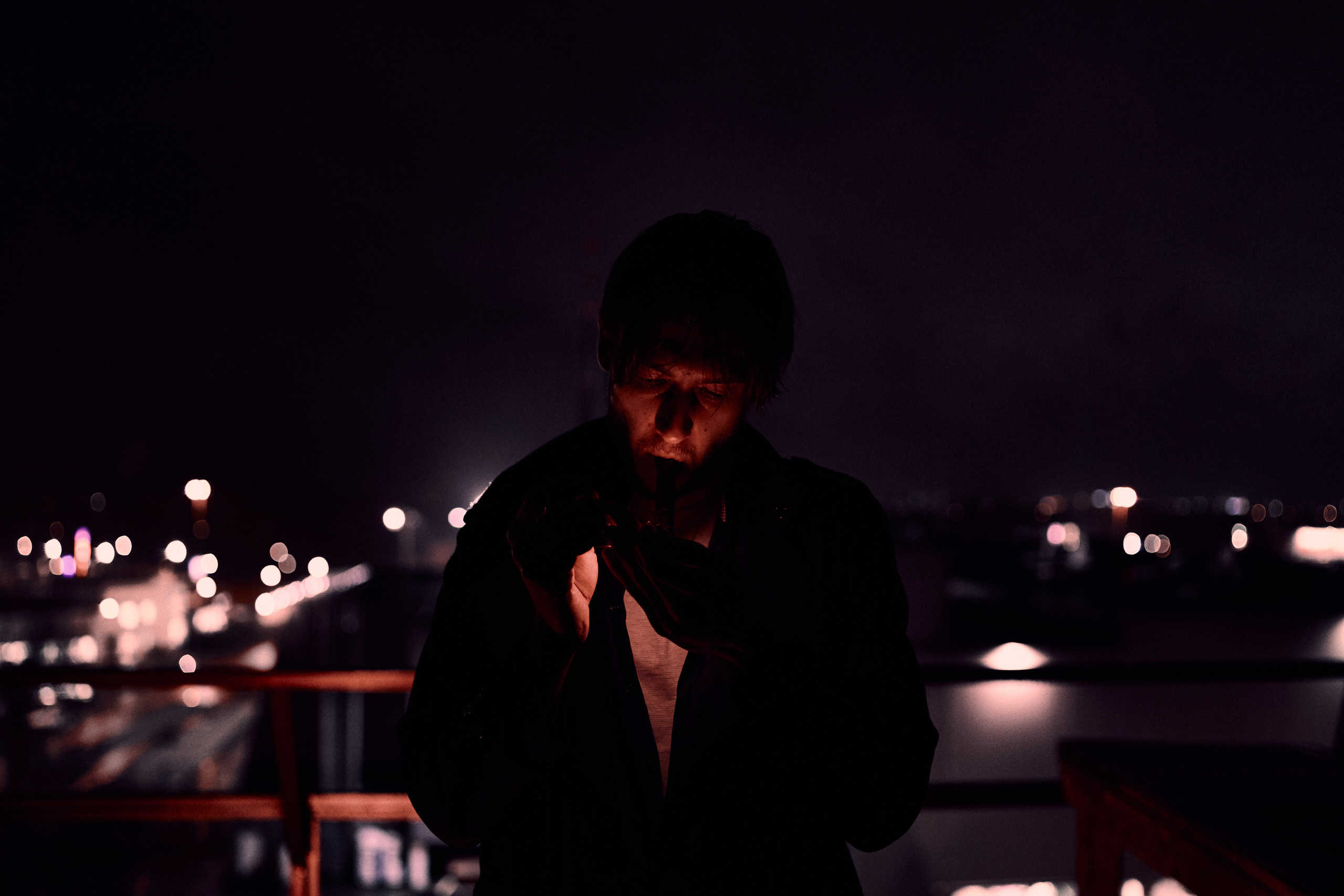A man standing on a rooftop at night, illuminated by soft light, with a city skyline blurred in the background, exuding a moody and reflective atmosphere.