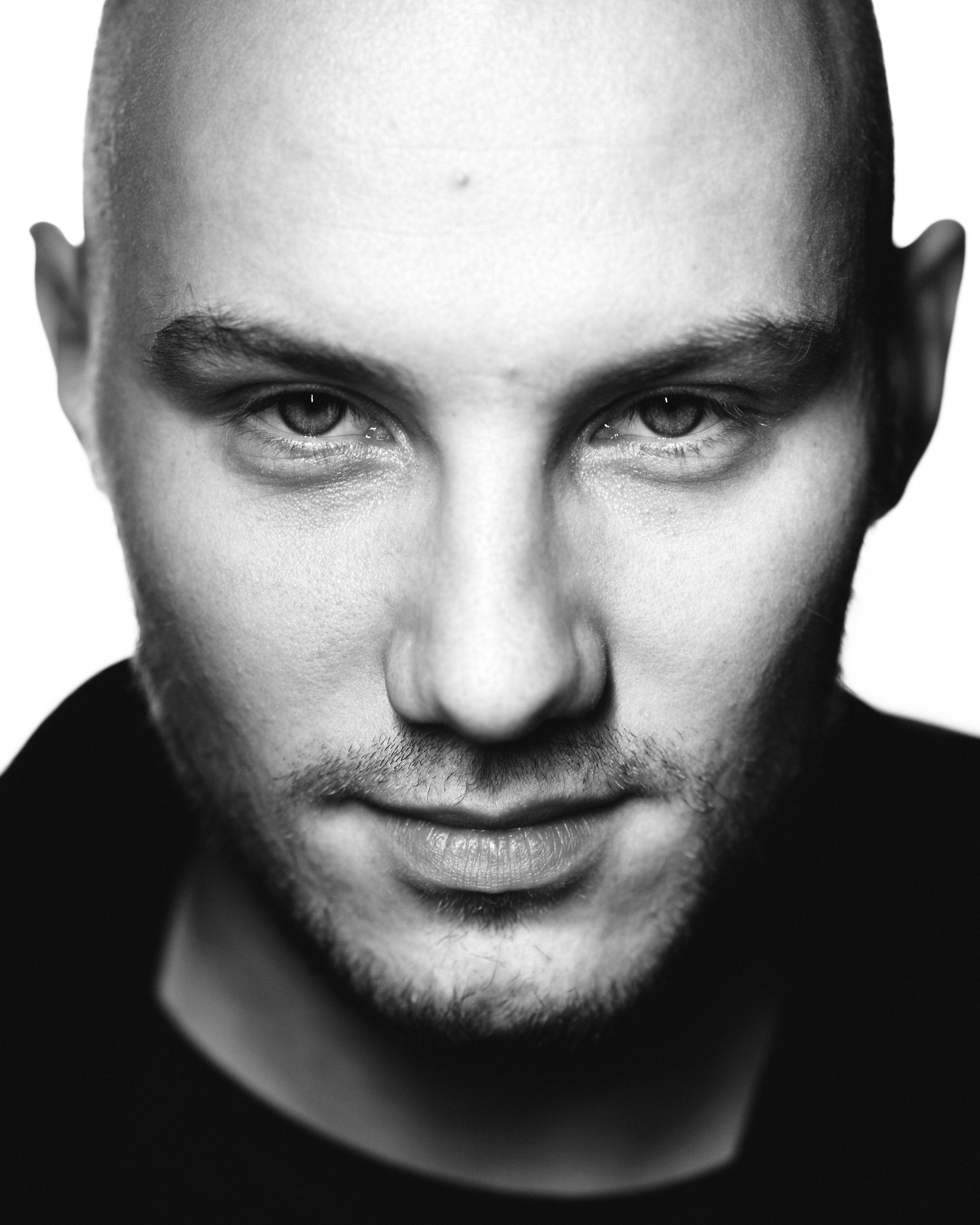 A striking black-and-white portrait of a man with a shaved head, staring directly at the camera with intensity.