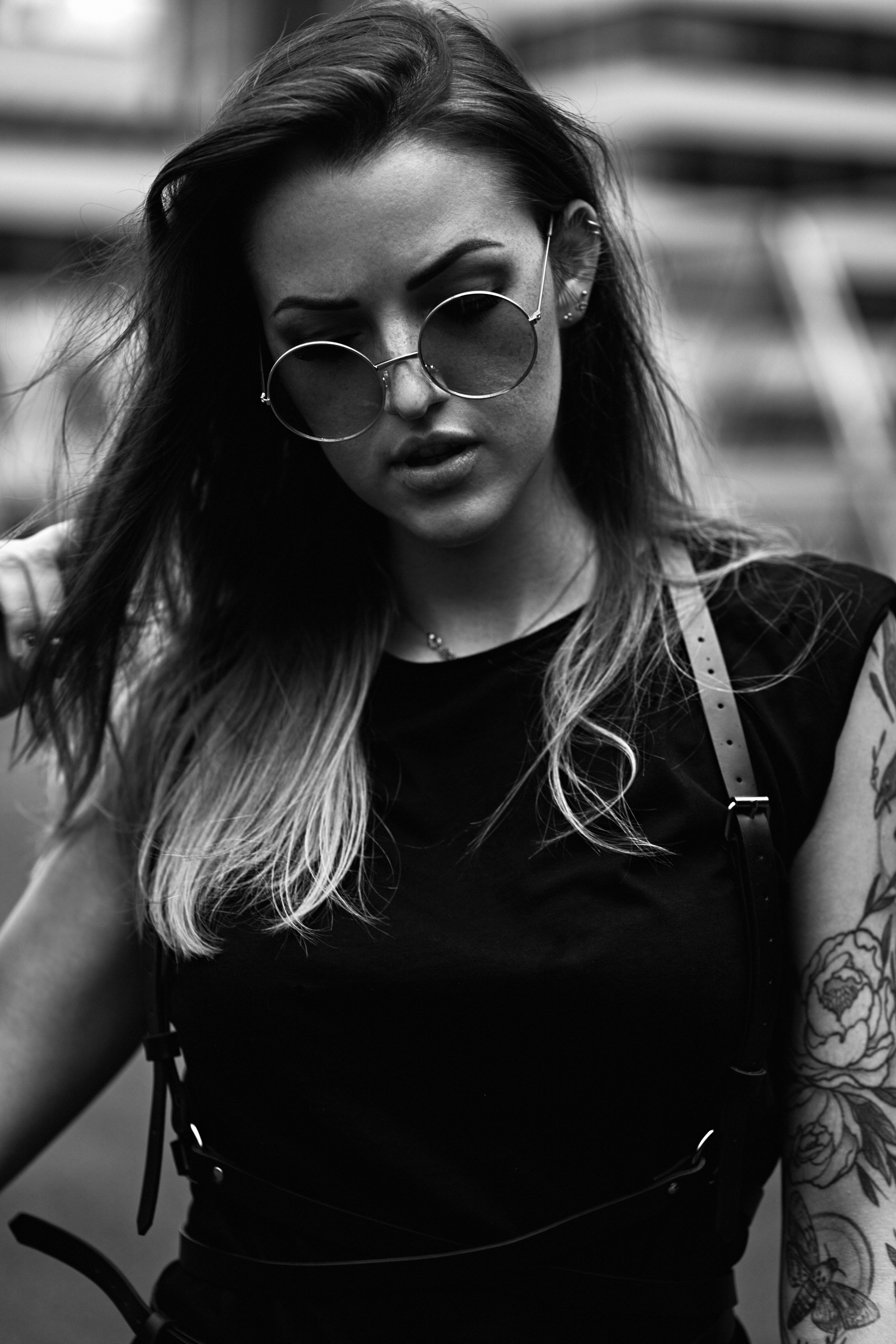 A portrait of a woman in sunglasses, her hair tousled, standing in an urban environment with a confident and edgy demeanor.