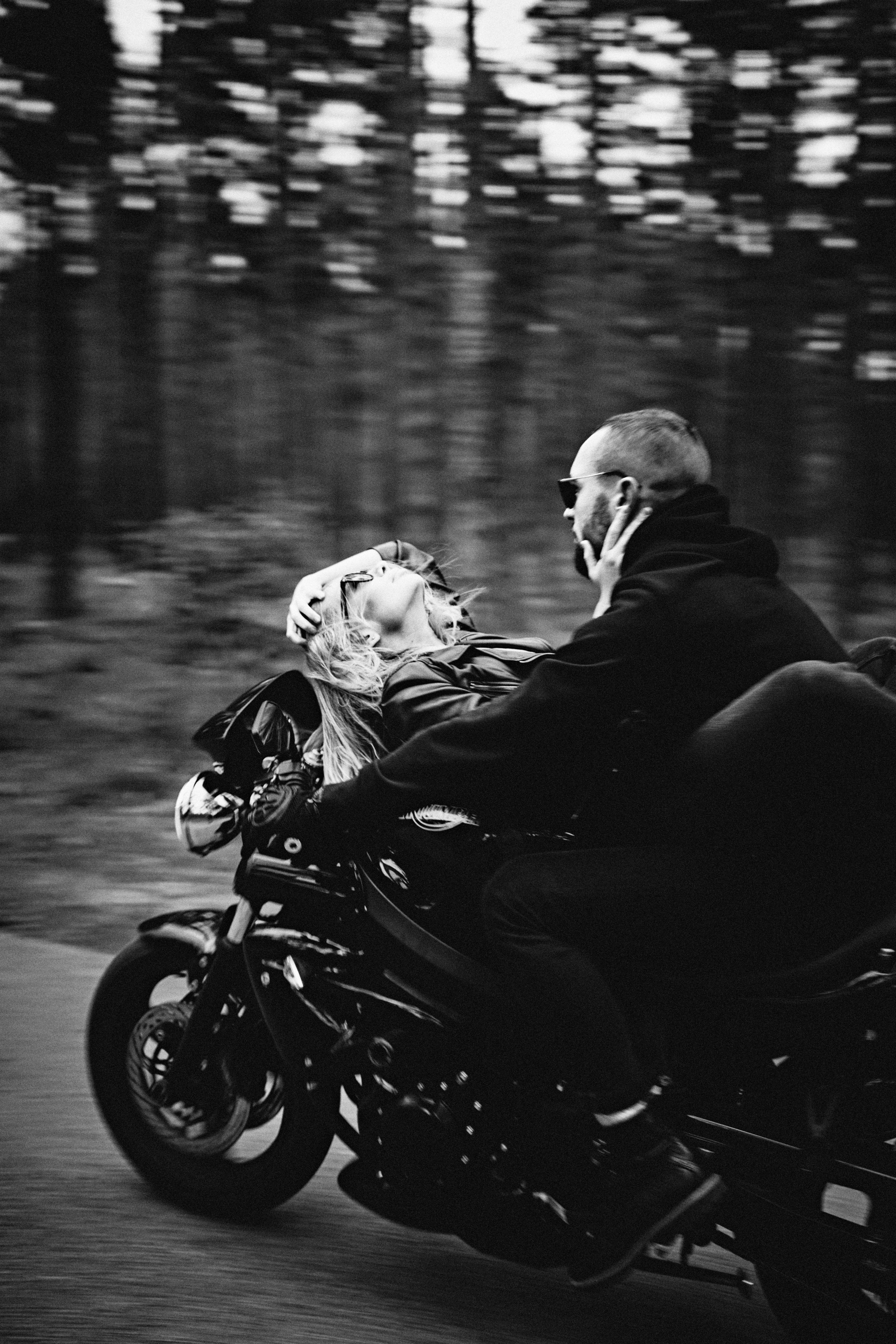 A woman lying on a motorcycle with a man driving, surrounded by blurred motion in a forested landscape.