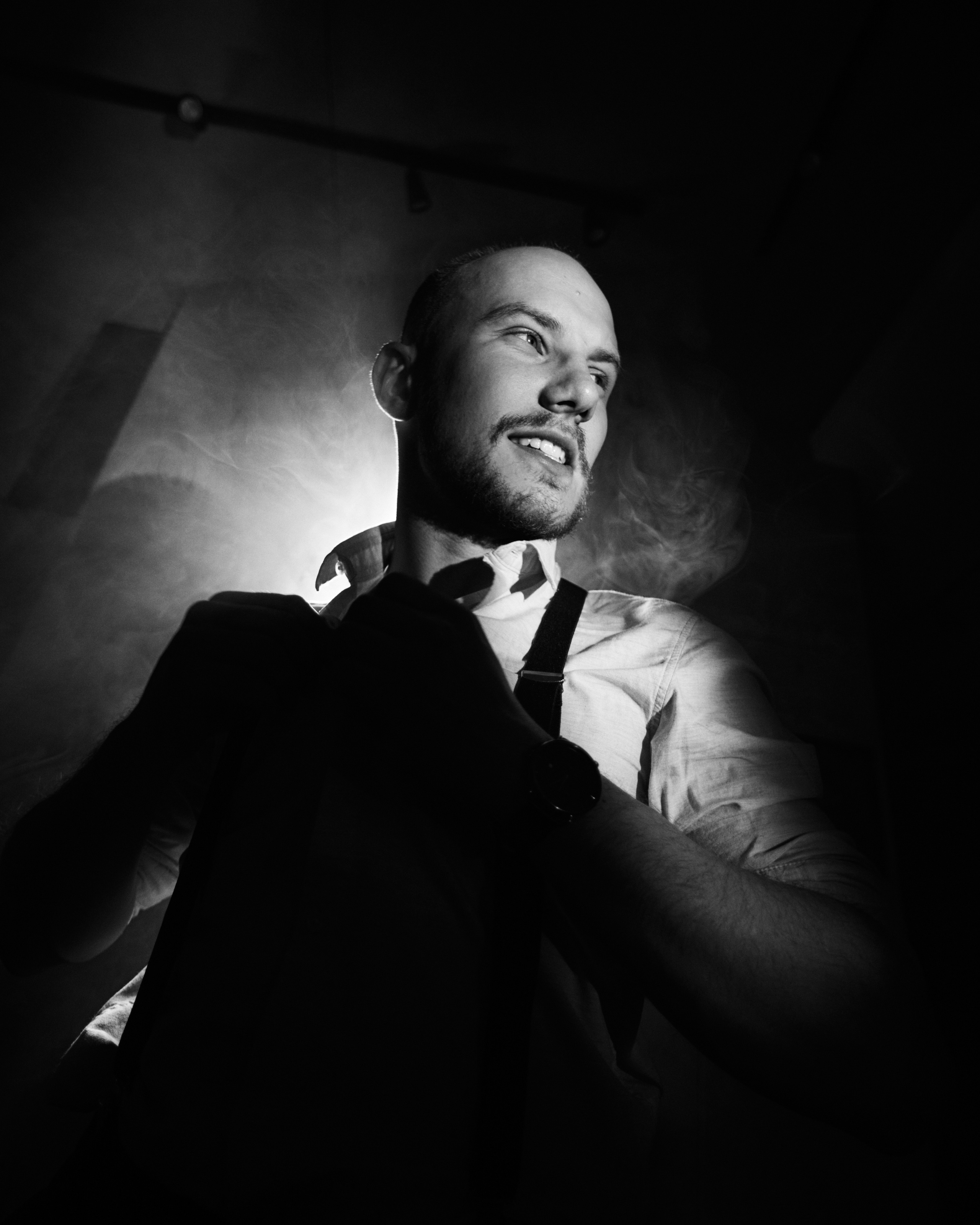 A man in a black-and-white studio portrait, adjusting his jacket, with dramatic lighting creating depth and character.