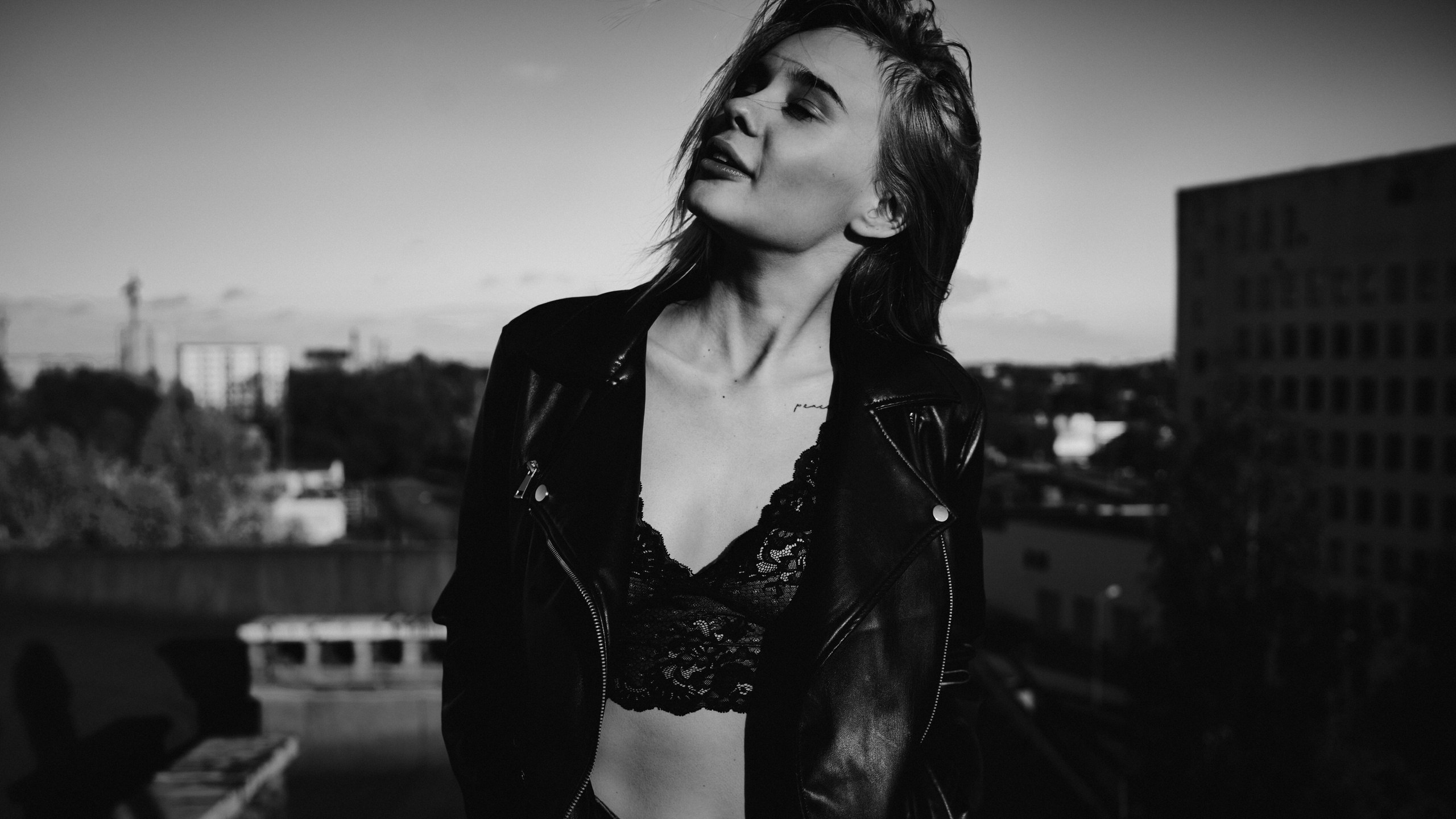 A  black-and-white portrait of a woman standing on a rooftop, wearing a lace top, with the city skyline in the background, exuding confidence and elegance.