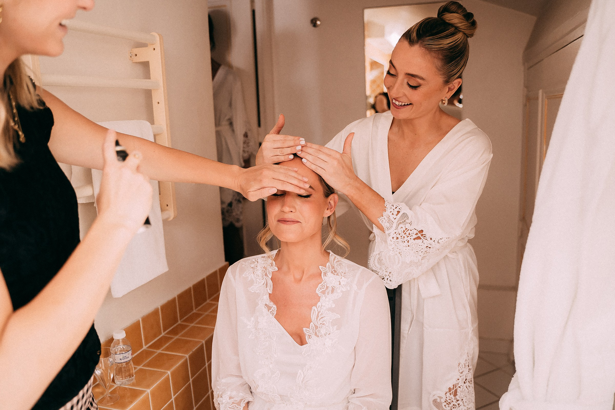 A bridesmaid lovingly adjusts the bride’s hair while preparing for the wedding in a warm and bright bathroom setting.