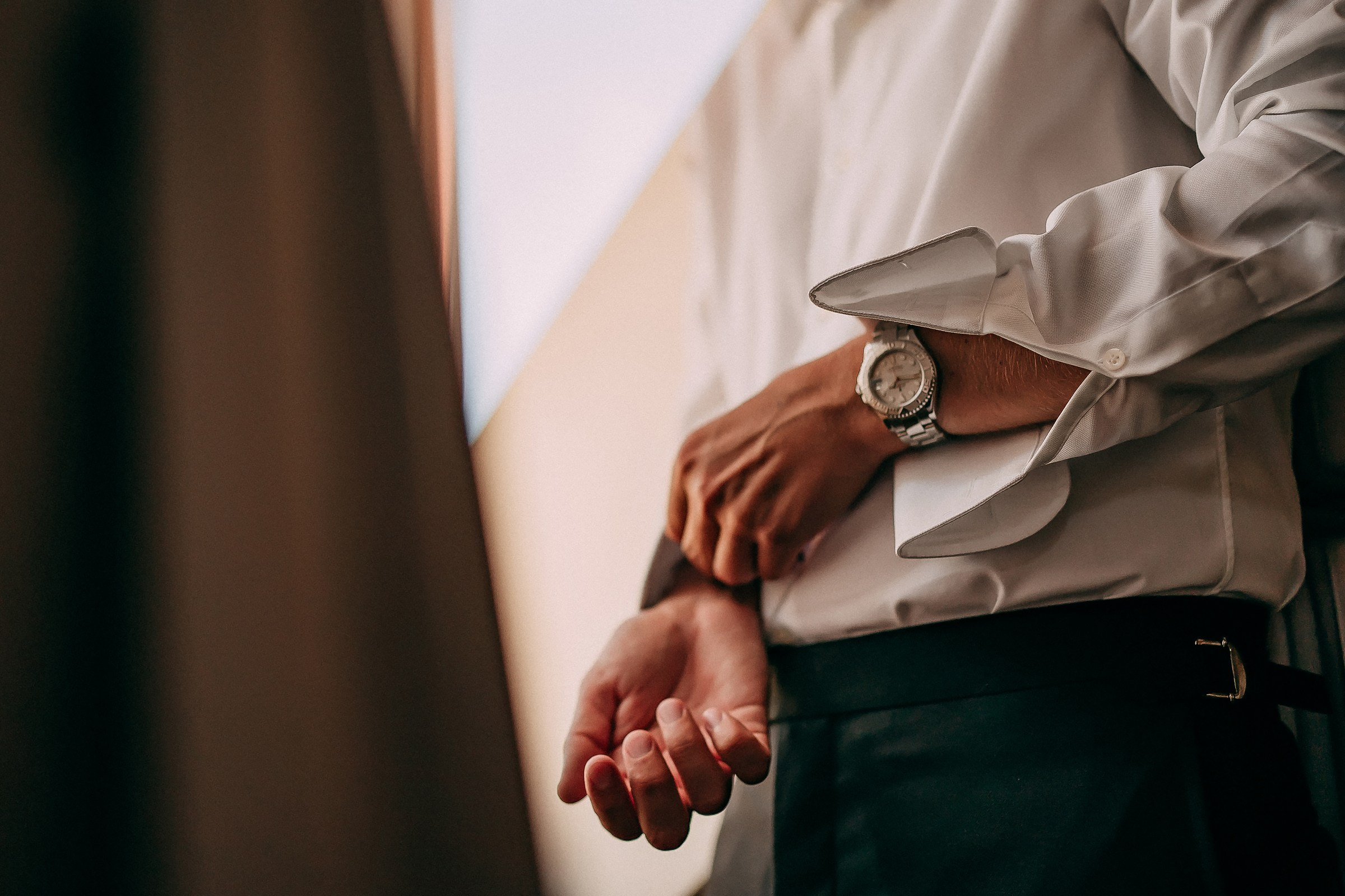 A close-up of a groom’s hands as he adjusts his cuff, wearing a stylish wristwatch. The image captures a tender and intimate moment before the wedding ceremony.